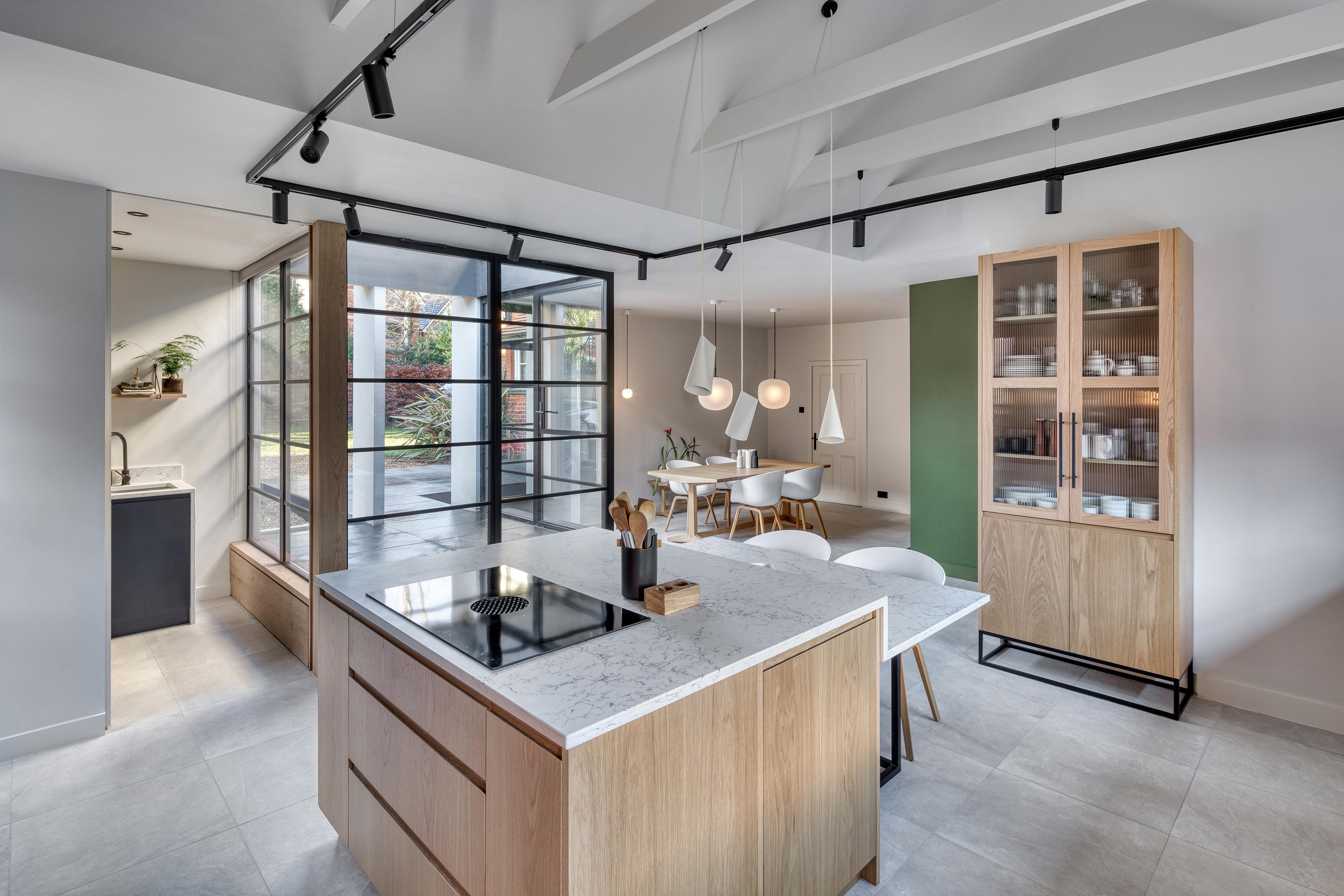 A modern kitchen and dining area with a marble island, wooden cabinets, and a built-in stovetop. The open layout features large windows, a green accent wall, and minimalist pendant lights over a dining table with white chairs
