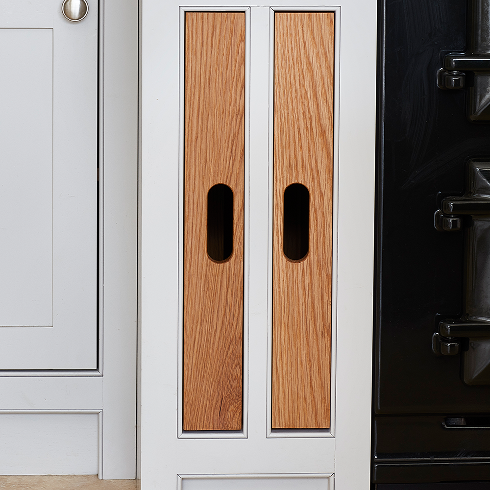 Two tall, narrow wooden cabinet doors with oval cut-out handles are set within a white kitchen unit, next to a white drawer and a black appliance.