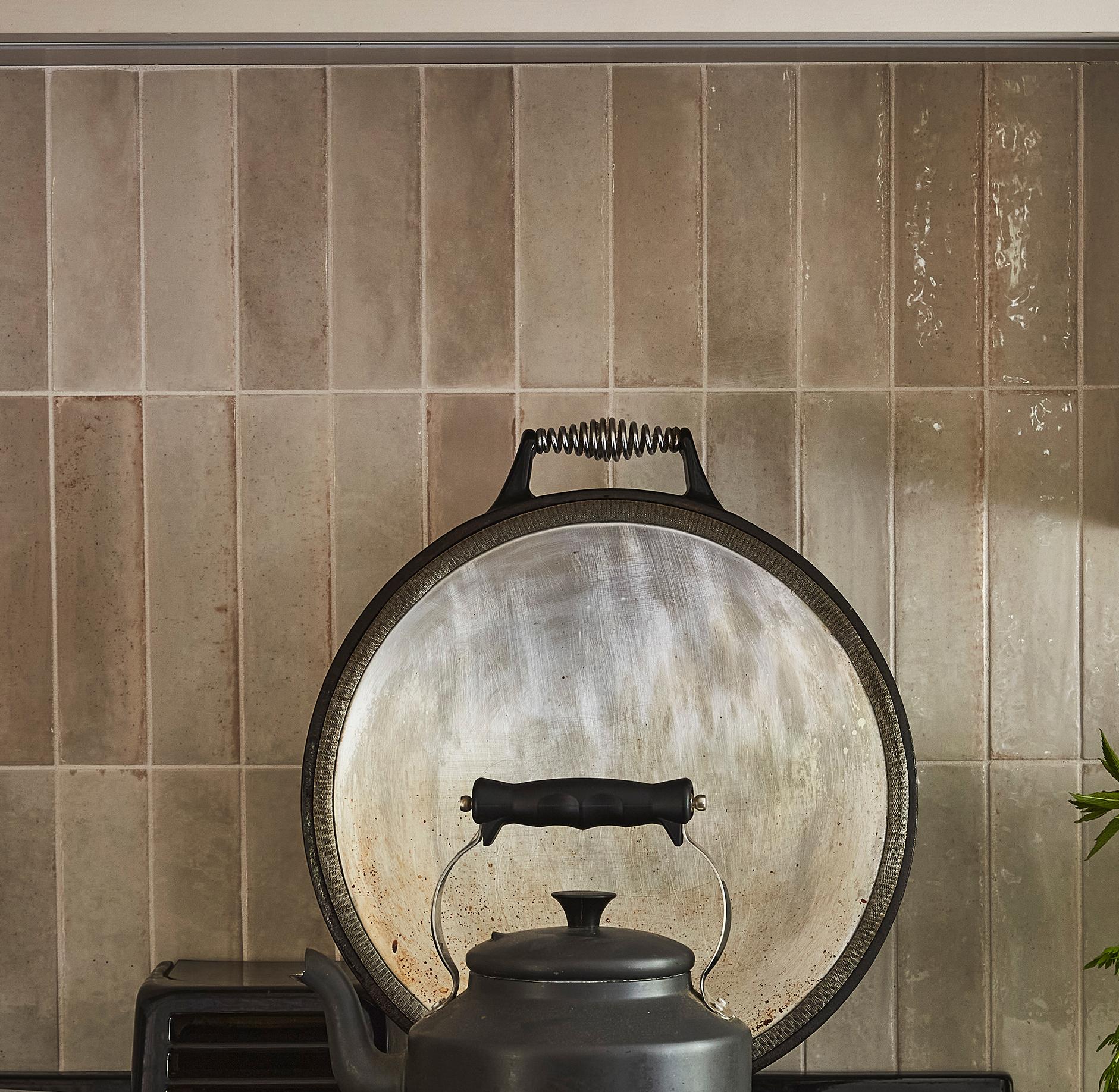 A black kettle with a curved handle sits on a stove in front of a large, round metal lid, against a beige tiled backsplash. Some green leaves are visible on the right edge of the image.