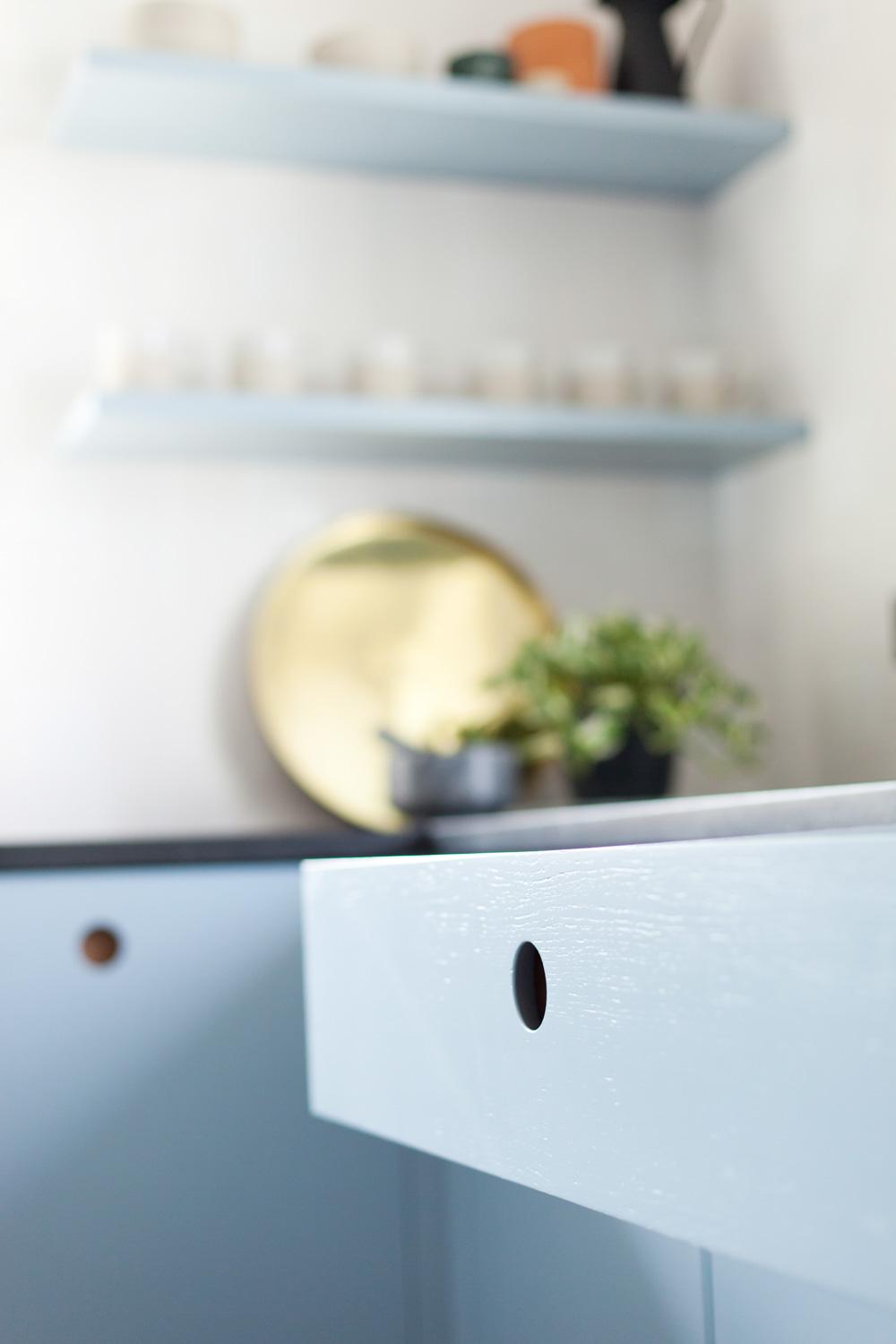 Close-up of a light blue kitchen drawer with a round hole handle. In the background, a gold tray and a small green plant are placed on a black countertop. Two shelves with small items are mounted on a white wall.