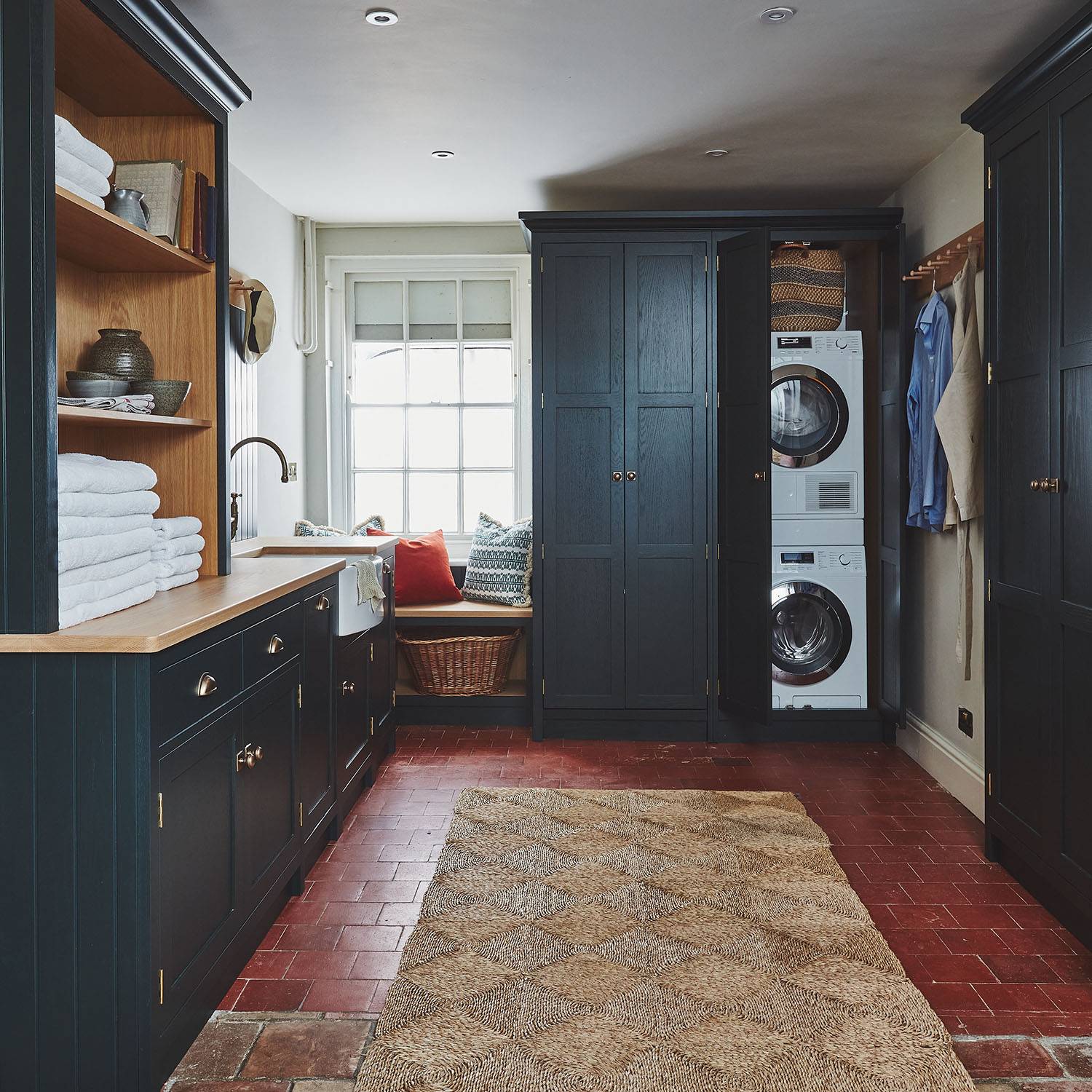 Laundry room with dark cabinets, a stacked washer and dryer, brick floor, and a woven rug. Wooden countertop holds folded towels. A window seat with cushions is in the corner, and there are hangers with shirts on the right.