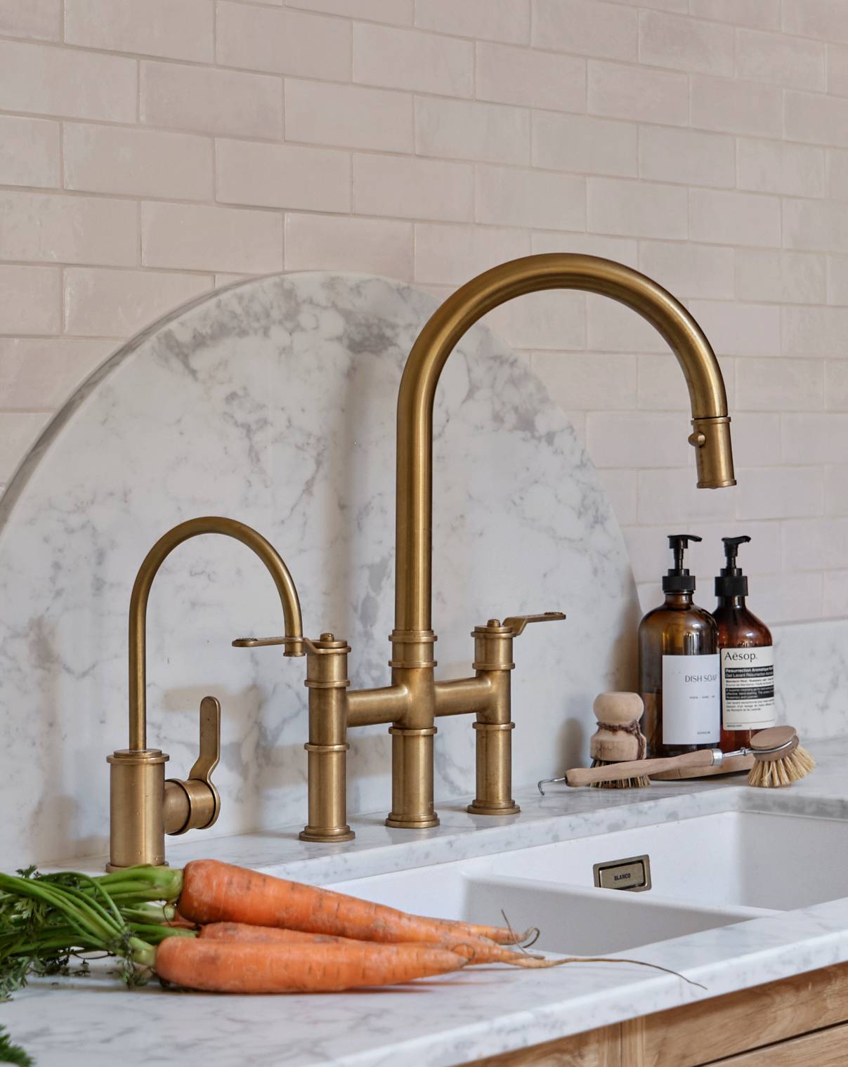A modern kitchen sink with brass faucets, white marble backsplash, and countertop. Fresh carrots, soap dispensers, and a dish brush are placed near the sink.
