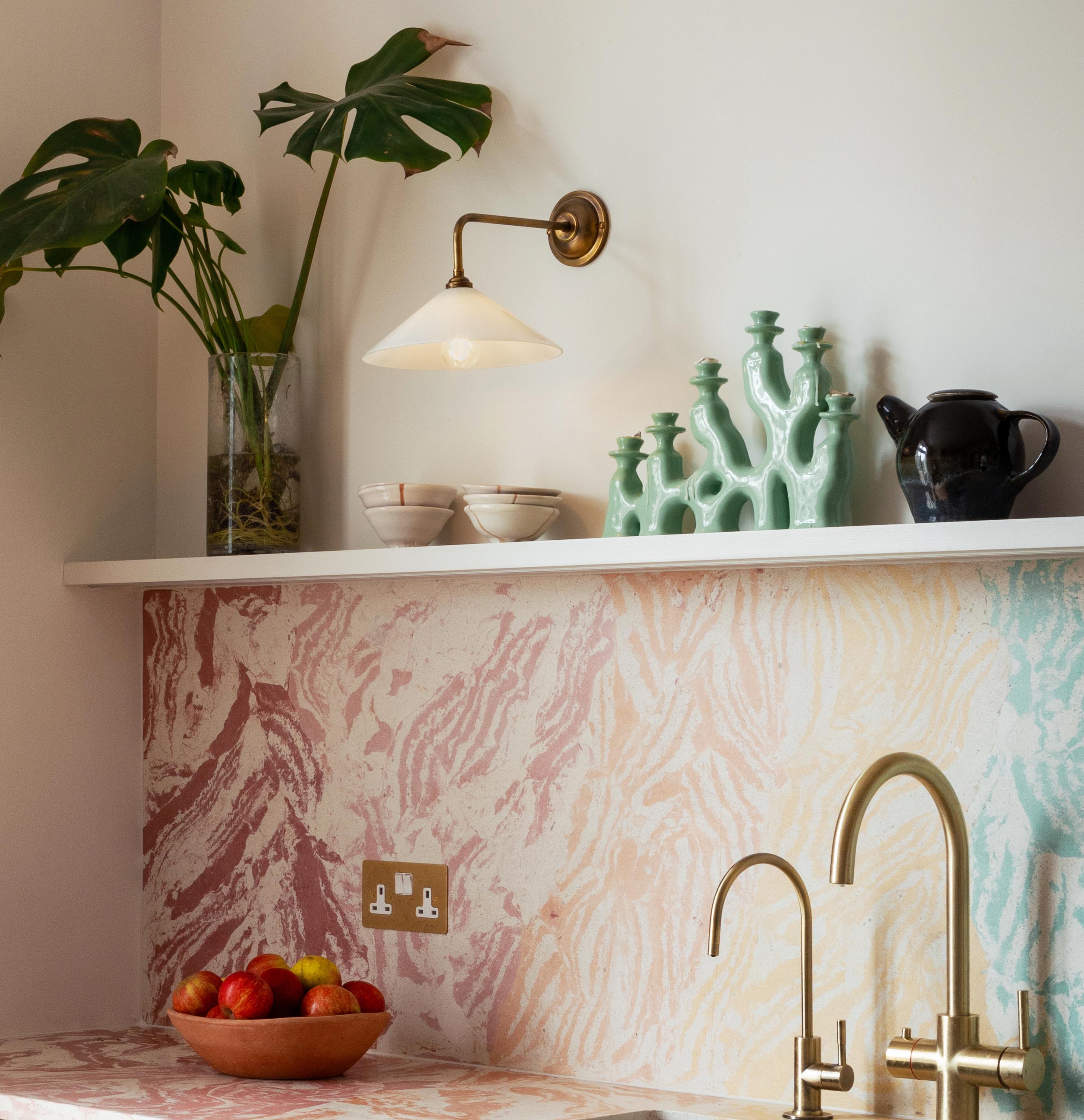 A kitchen corner with a colorful marbled countertop and backsplash. A brass faucet is mounted below a wall shelf. The shelf holds a vase with Monstera leaves, bowls, a teapot, and ceramic decor. A wall sconce provides light. A bowl of fruit is on the counter.
