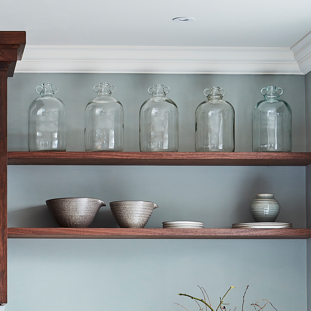 Two wooden shelves on a gray wall hold five large clear glass jars on the top shelf and a set of ceramic bowls and plates on the lower shelf, creating a minimalist and tidy kitchen display