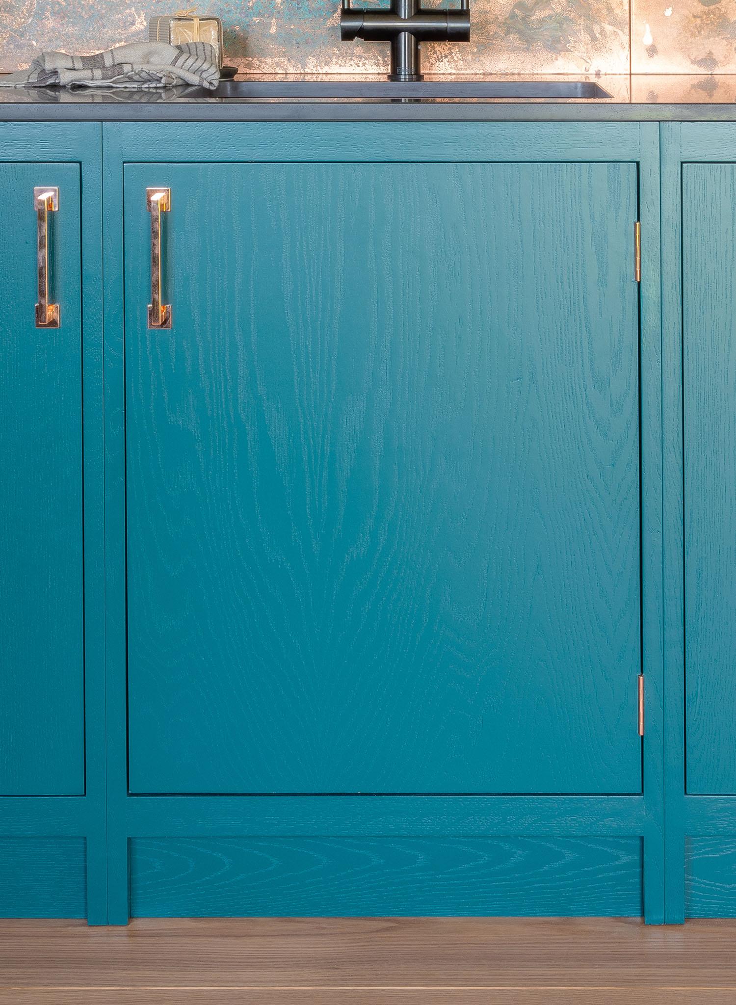 Teal kitchen cabinets with gold handles, a marble or stone backsplash, and a black faucet above the sink, with a towel and soap on the counter. Wood flooring is visible at the bottom of the image.