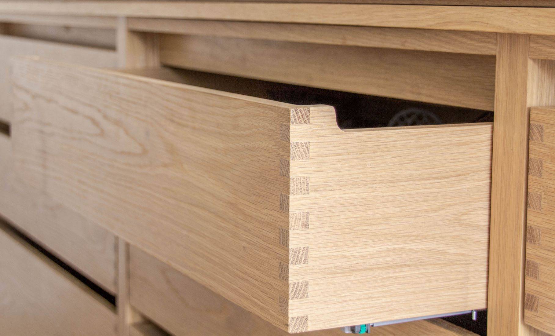 A close-up of a partially open wooden drawer with visible dovetail joints, showing the craftsmanship and smooth wood grain of the light-colored cabinet.