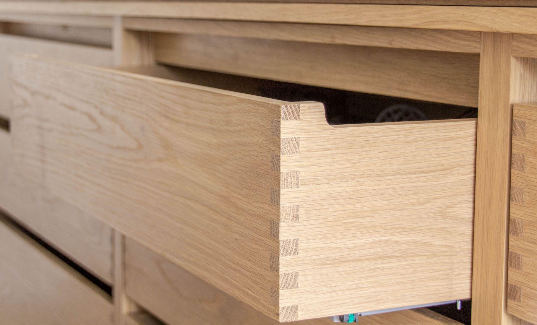 A close-up of a partially open wooden drawer with visible dovetail joints, showing the craftsmanship and smooth wood grain of the light-colored cabinet.