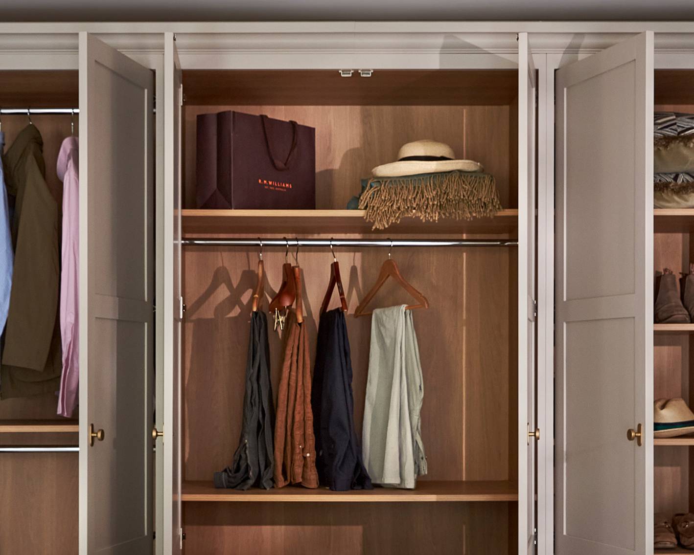 An organized wooden wardrobe with open doors displays hanging clothes, scarves on hooks, a brown shopping bag, a hat with a scarf, and shelves with neatly folded items.