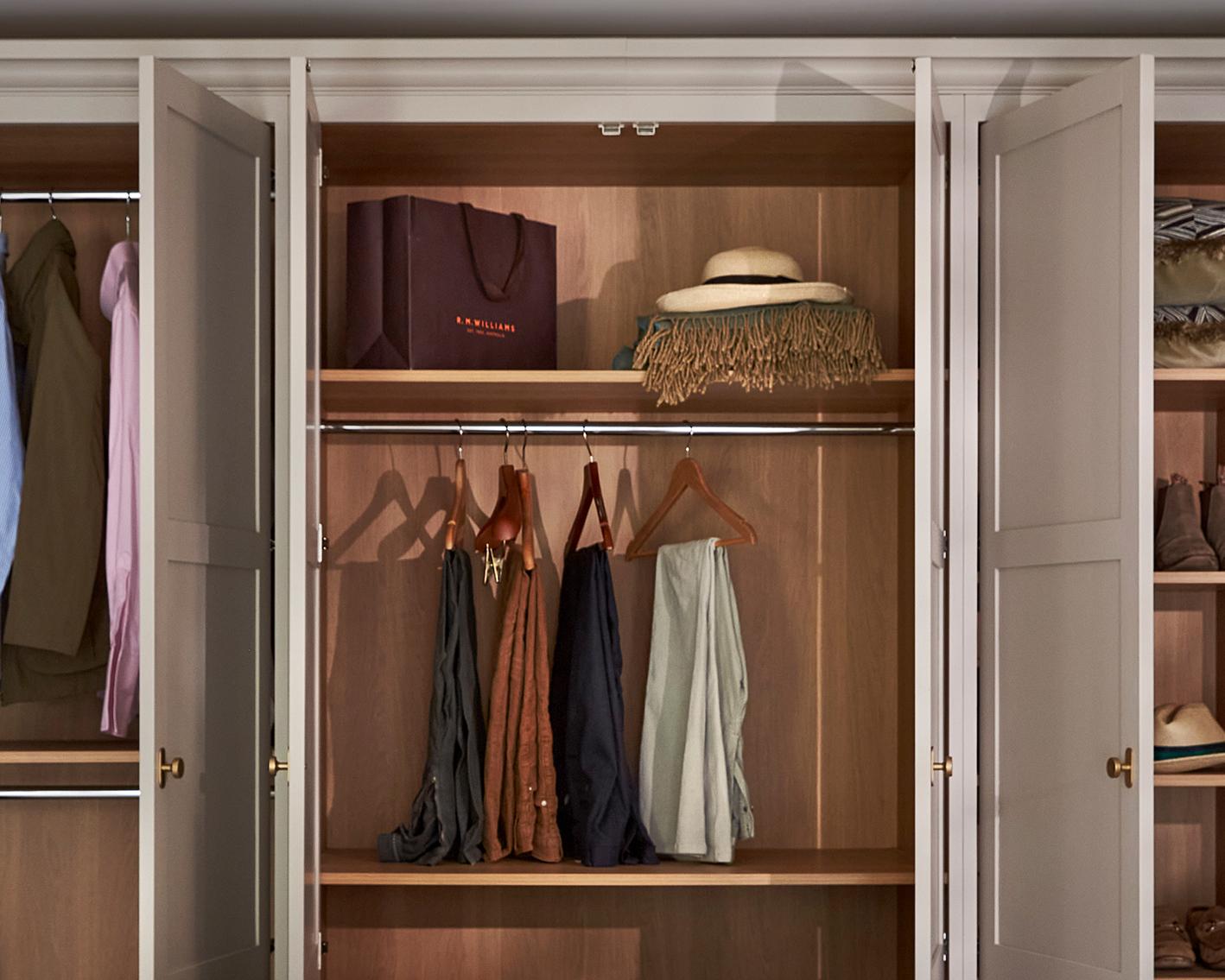 An organized wooden wardrobe with open doors displays hanging clothes, scarves on hooks, a brown shopping bag, a hat with a scarf, and shelves with neatly folded items.