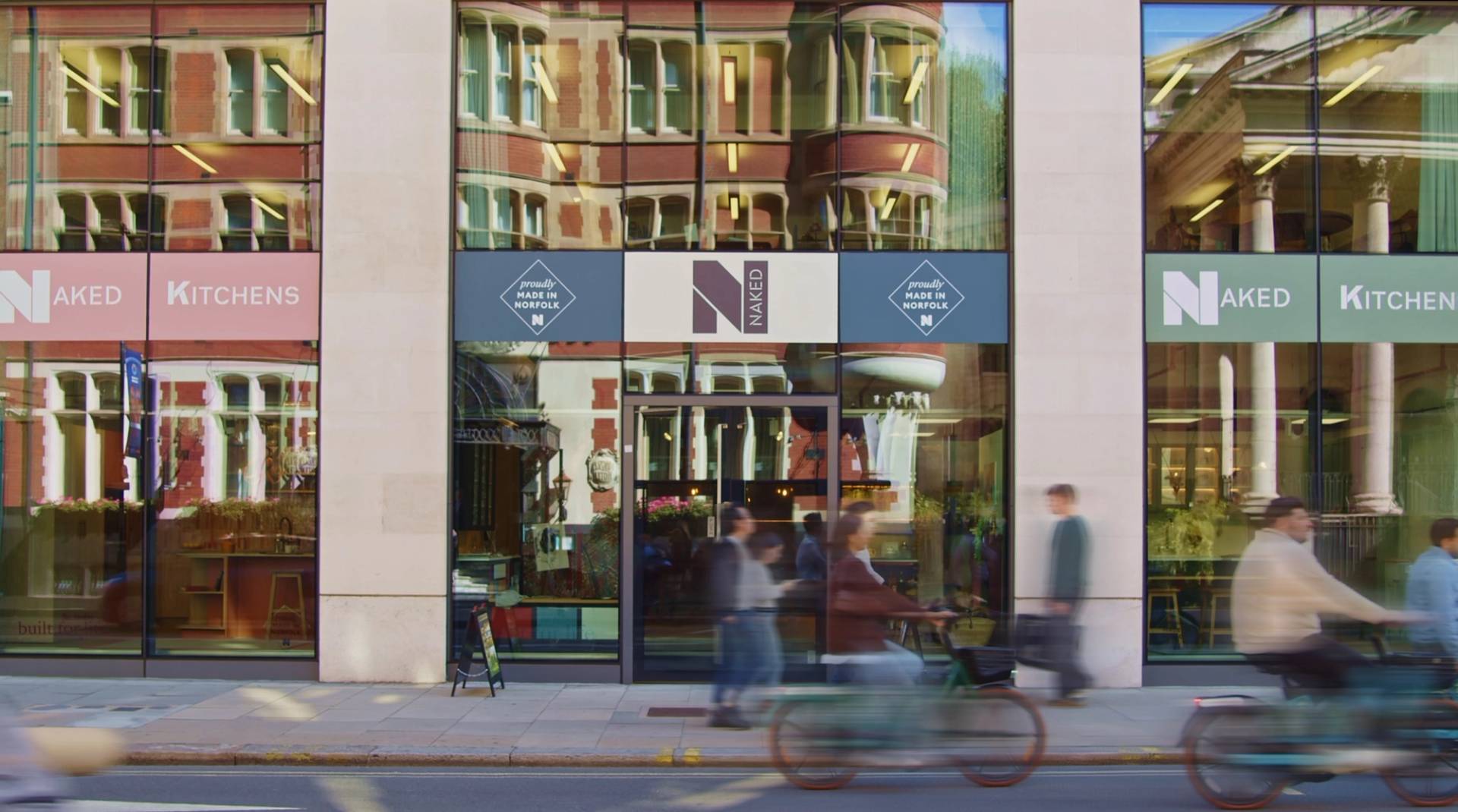 A storefront with large glass windows reflecting a brick building. A sign with 'NAKED' is displayed above the door. People walk by on the sidewalk, and a cyclist rides past the shop.
