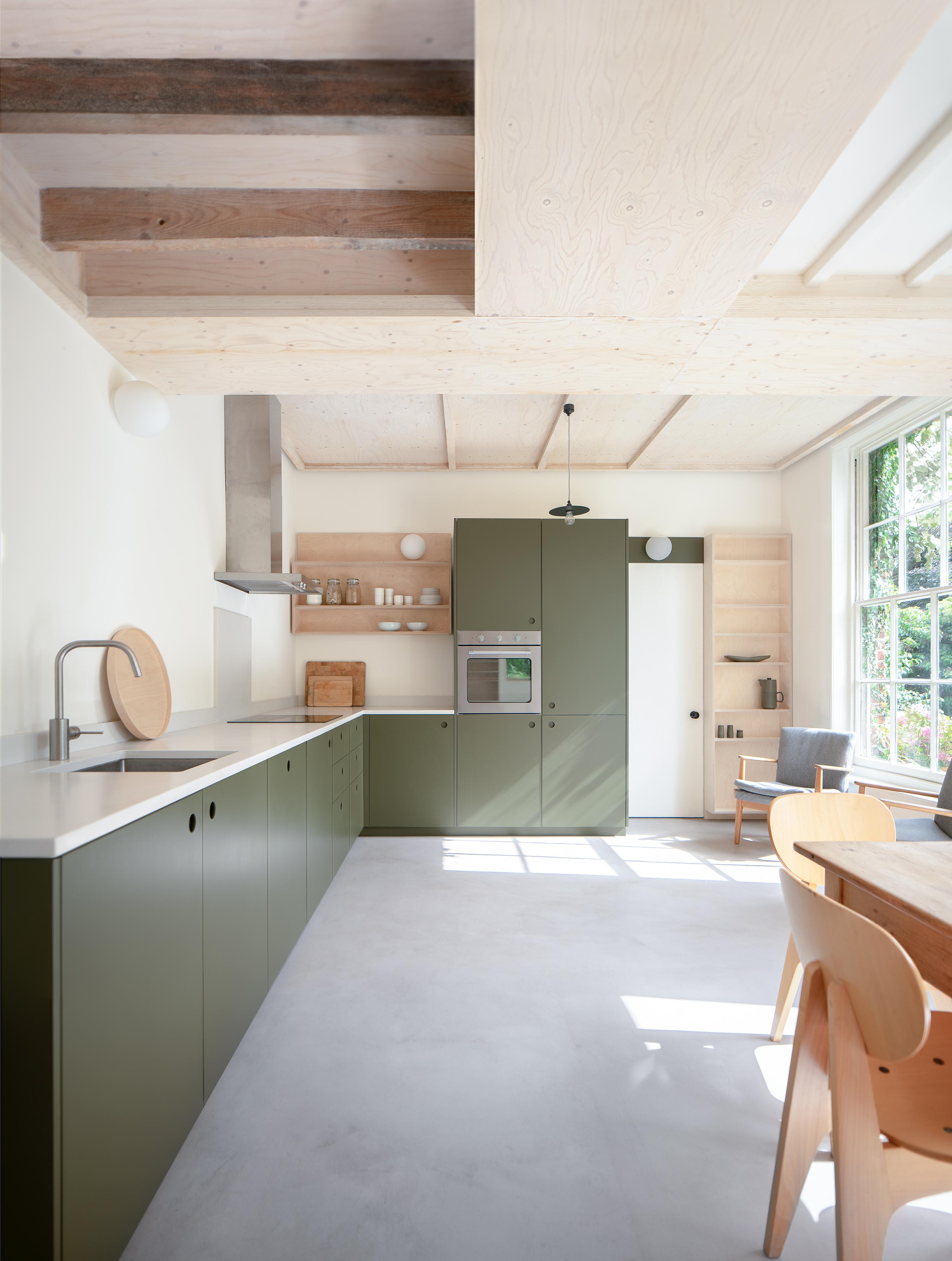 A modern kitchen with olive green cabinets, white countertops, and a built-in oven. Open shelving displays kitchenware. A light wooden dining table with chairs is on the right, and large windows let in natural light. Ceiling features exposed wood beams.