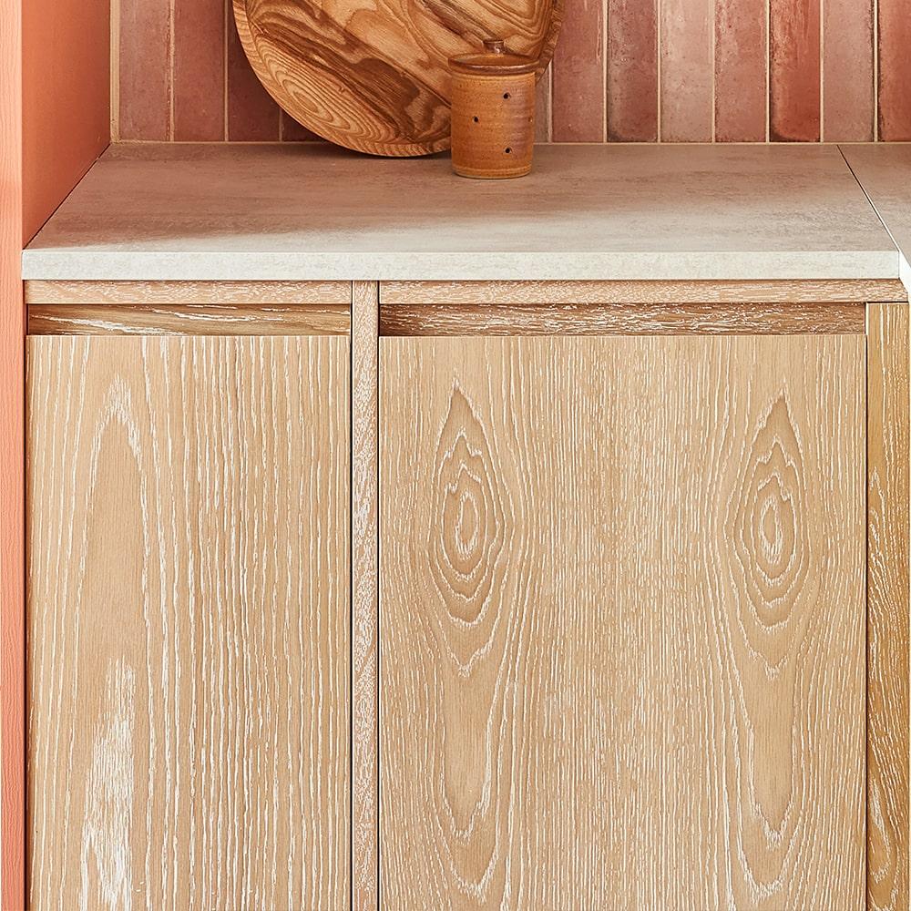 Light wood grain cabinets with a white countertop and vertical pink tiles on the wall. A wooden cutting board and small container are placed on the counter for decoration.