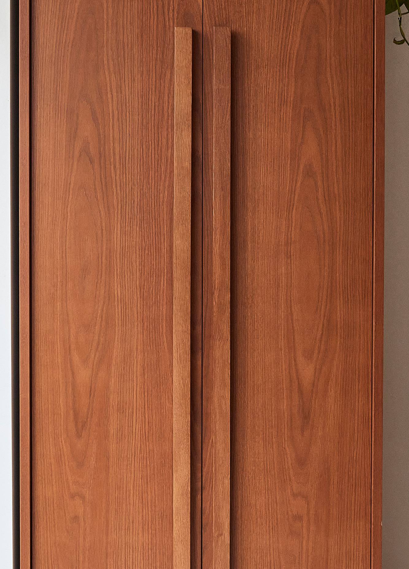 Wooden cabinet doors with vertical grain patterns, featuring tall, slim handles. The warm brown tone adds a classic touch, with a hint of green plant visible at the top right corner.