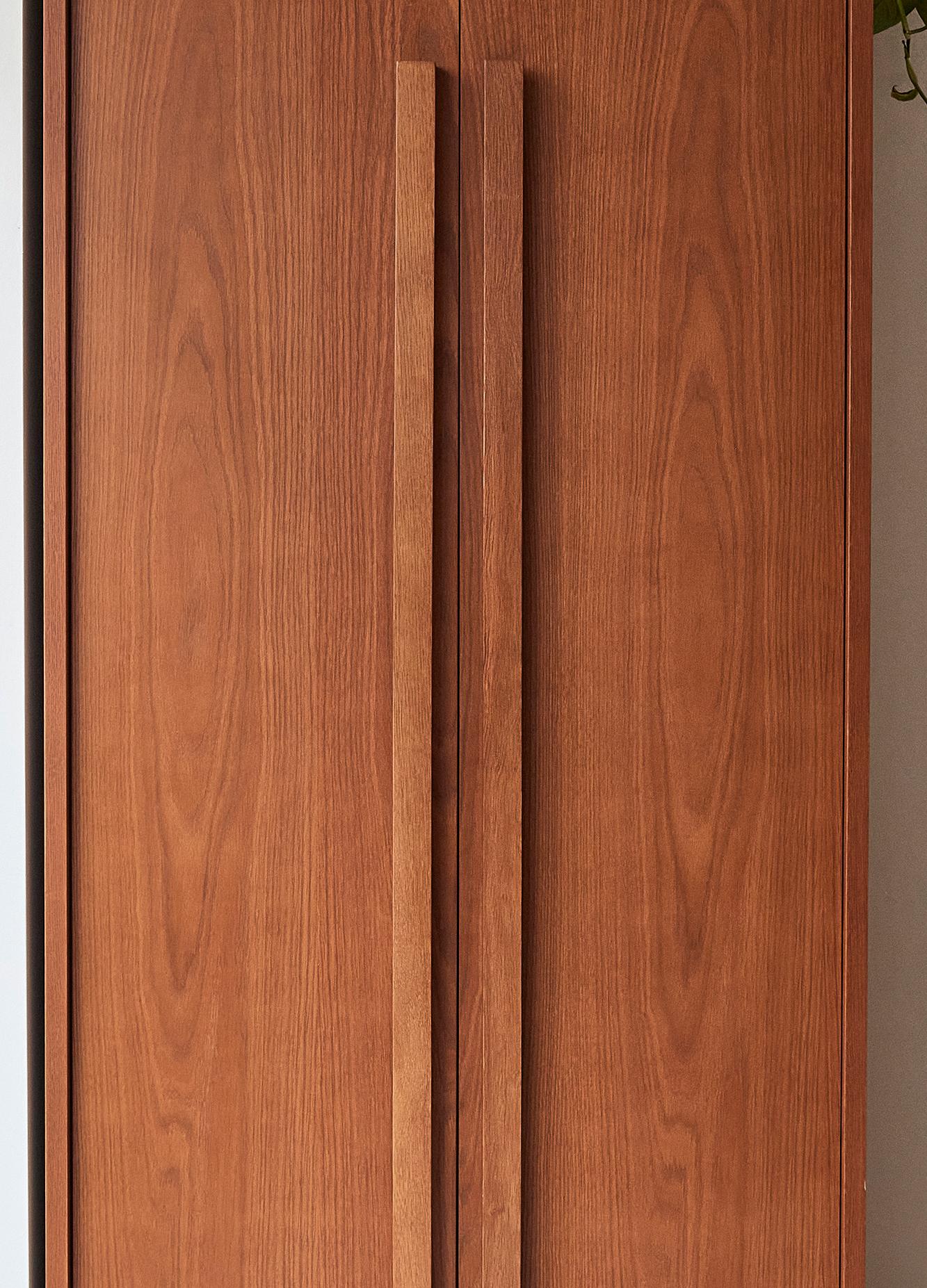 Wooden cabinet doors with vertical grain patterns, featuring tall, slim handles. The warm brown tone adds a classic touch, with a hint of green plant visible at the top right corner.