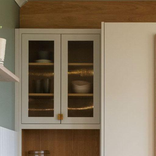 A modern kitchen cabinet with frosted glass doors displaying dishes and a bowl on two shelves. The cabinet is white with wooden accents and gold handles, complementing the green and cream wall colors and a shelf with stacked items.