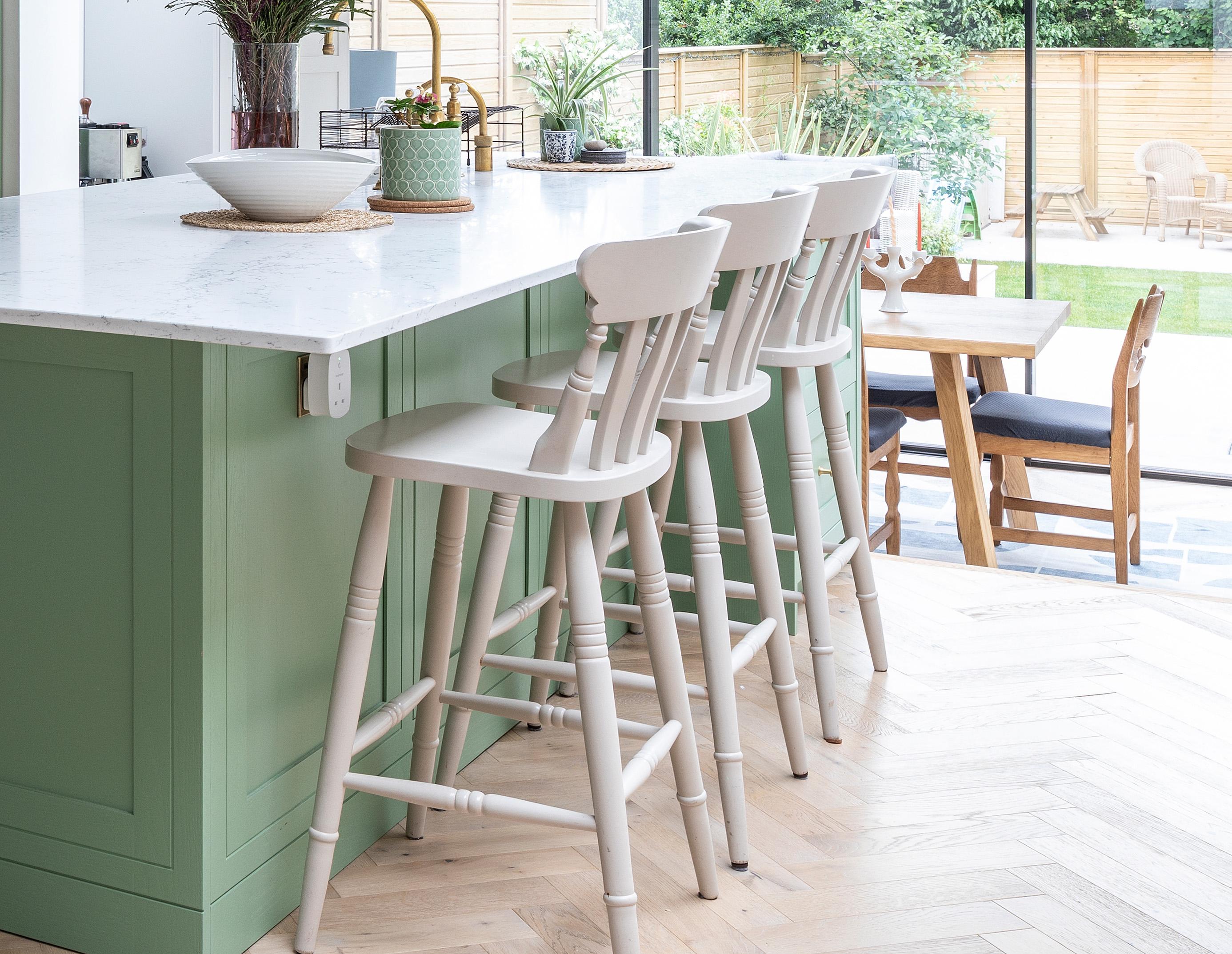 Four white wooden bar stools line a light green kitchen island with a white countertop in a bright, modern kitchen. In the background, there is a dining table and large windows overlooking a garden.