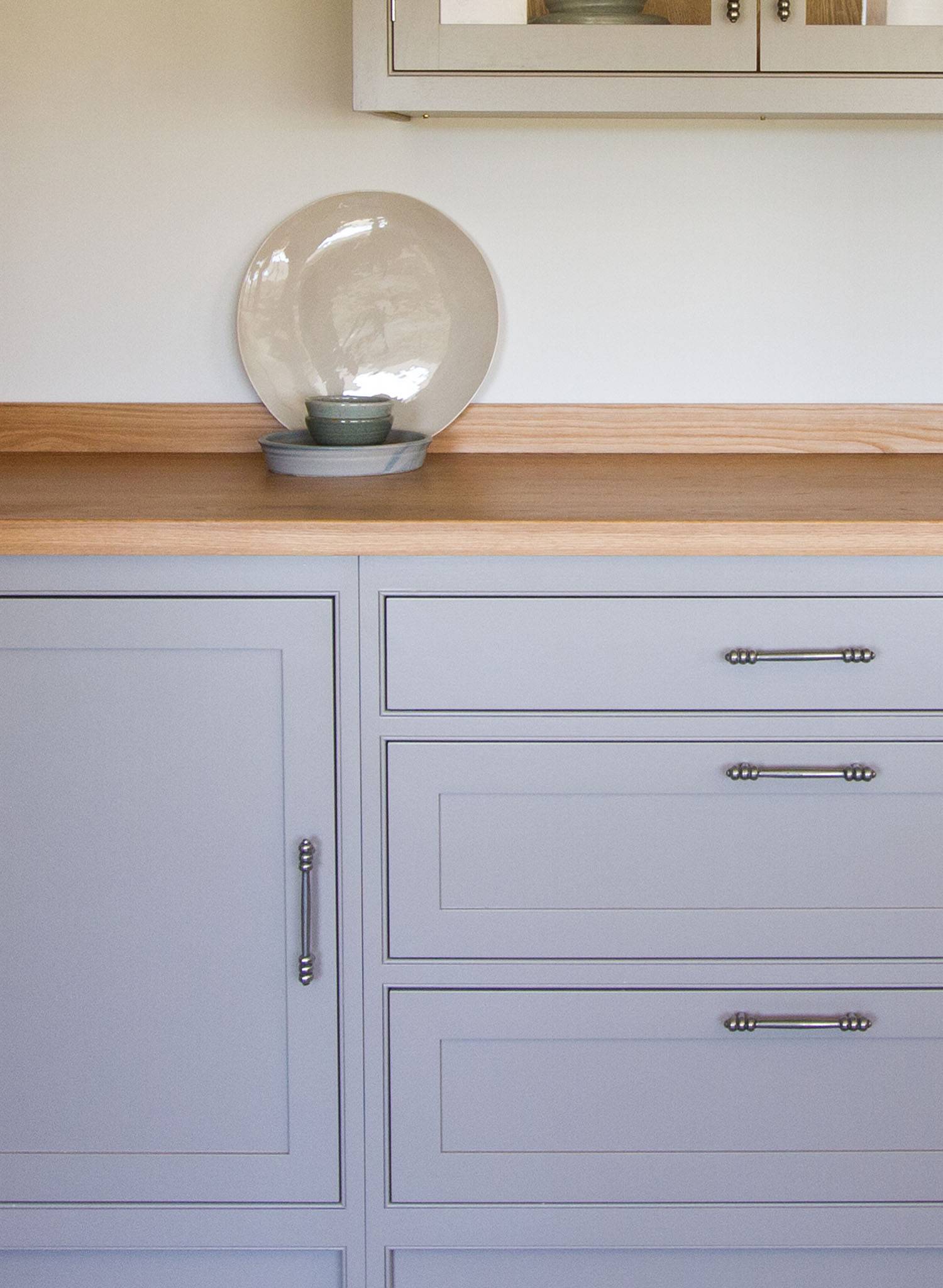 Light gray cabinets and drawers with silver handles, topped with a light wood countertop. On the counter sits a large round ceramic plate with a small bowl in front, both in neutral tones, against a plain white wall.