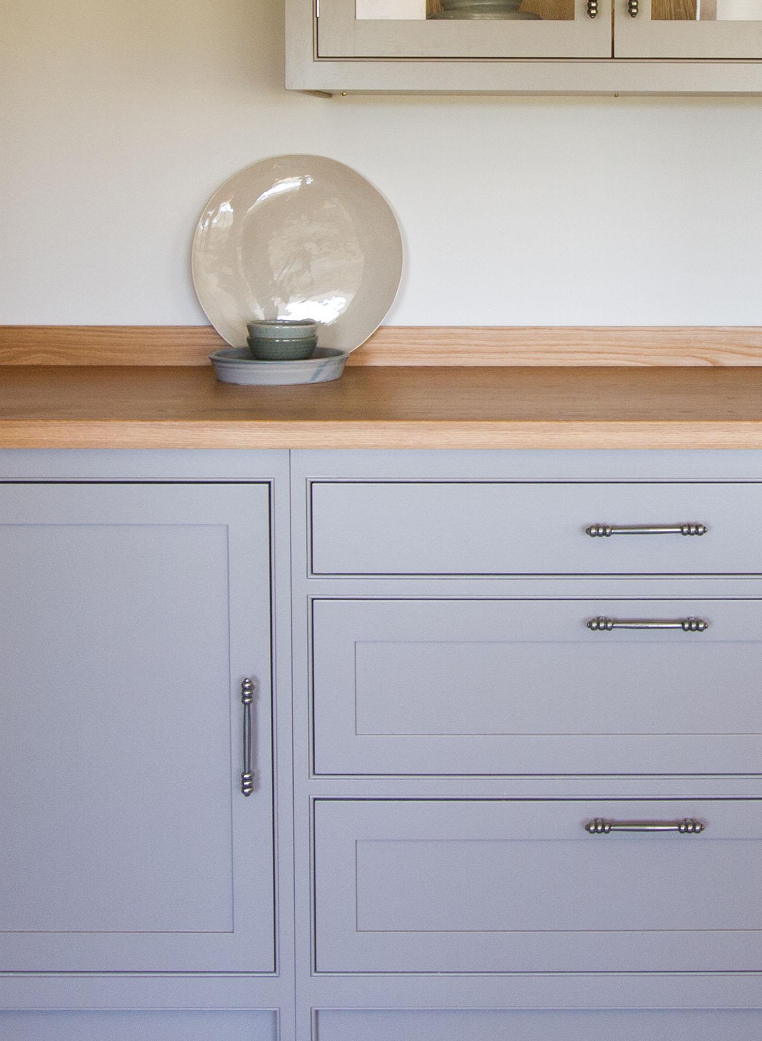 Light gray cabinets and drawers with silver handles, topped with a light wood countertop. On the counter sits a large round ceramic plate with a small bowl in front, both in neutral tones, against a plain white wall.