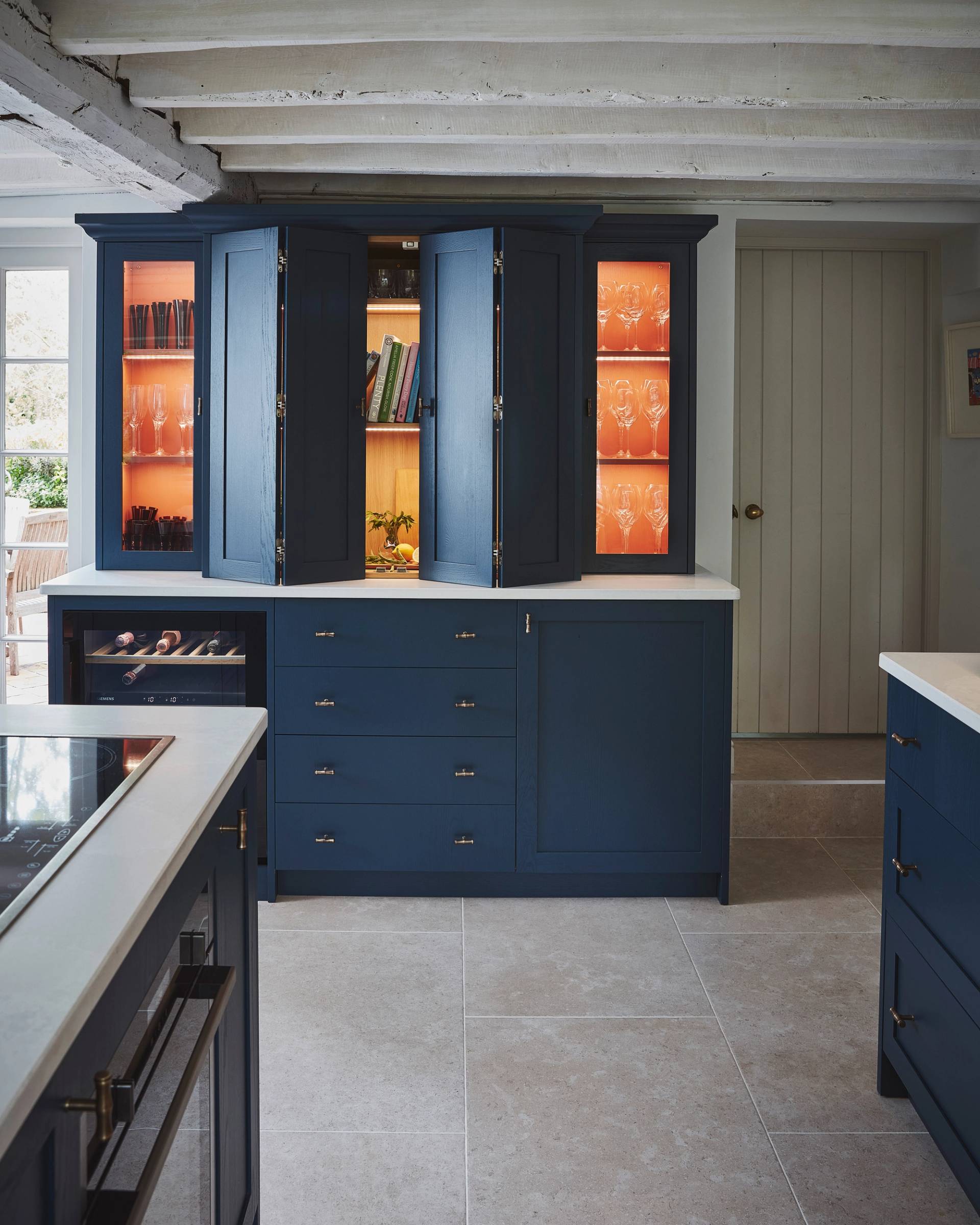 A blue kitchen cabinet with open doors reveals glassware, cookbooks, and a cutting board with a knife and sliced lemon. French doors lead out to a patio with wooden chairs and a table, surrounded by greenery.
