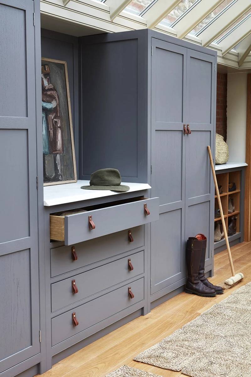 A stylish mudroom with gray cabinets, a drawer slightly open, and a shelf displaying a hat and a figurative painting. A pair of boots and a polo mallet are on the wooden floor near a beige rug. Natural light streams through windows above.