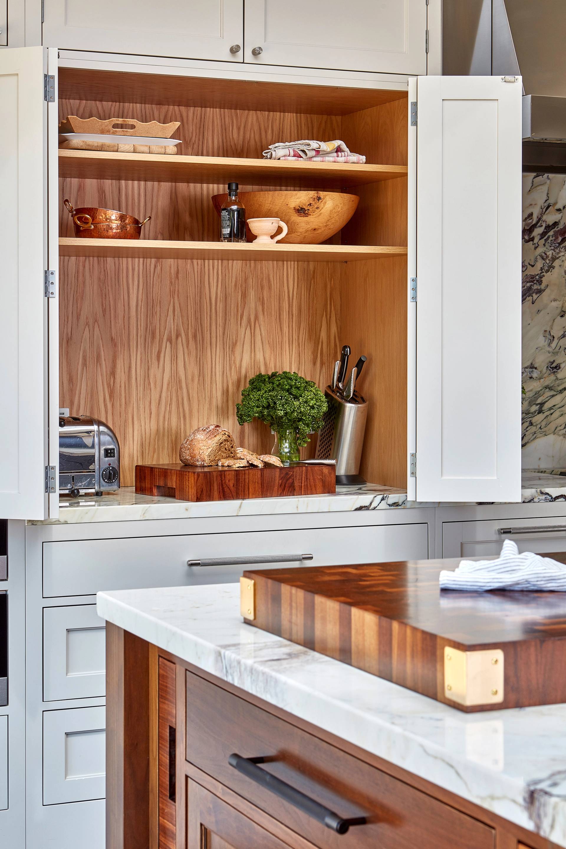 A kitchen with white cabinets and a natural wood interior. Shelves display a copper pot, bowls, a pepper grinder, and a bottle. A chopping block holds parsley and a bread loaf. Nearby are a toaster and knife block. Marble countertops add elegance