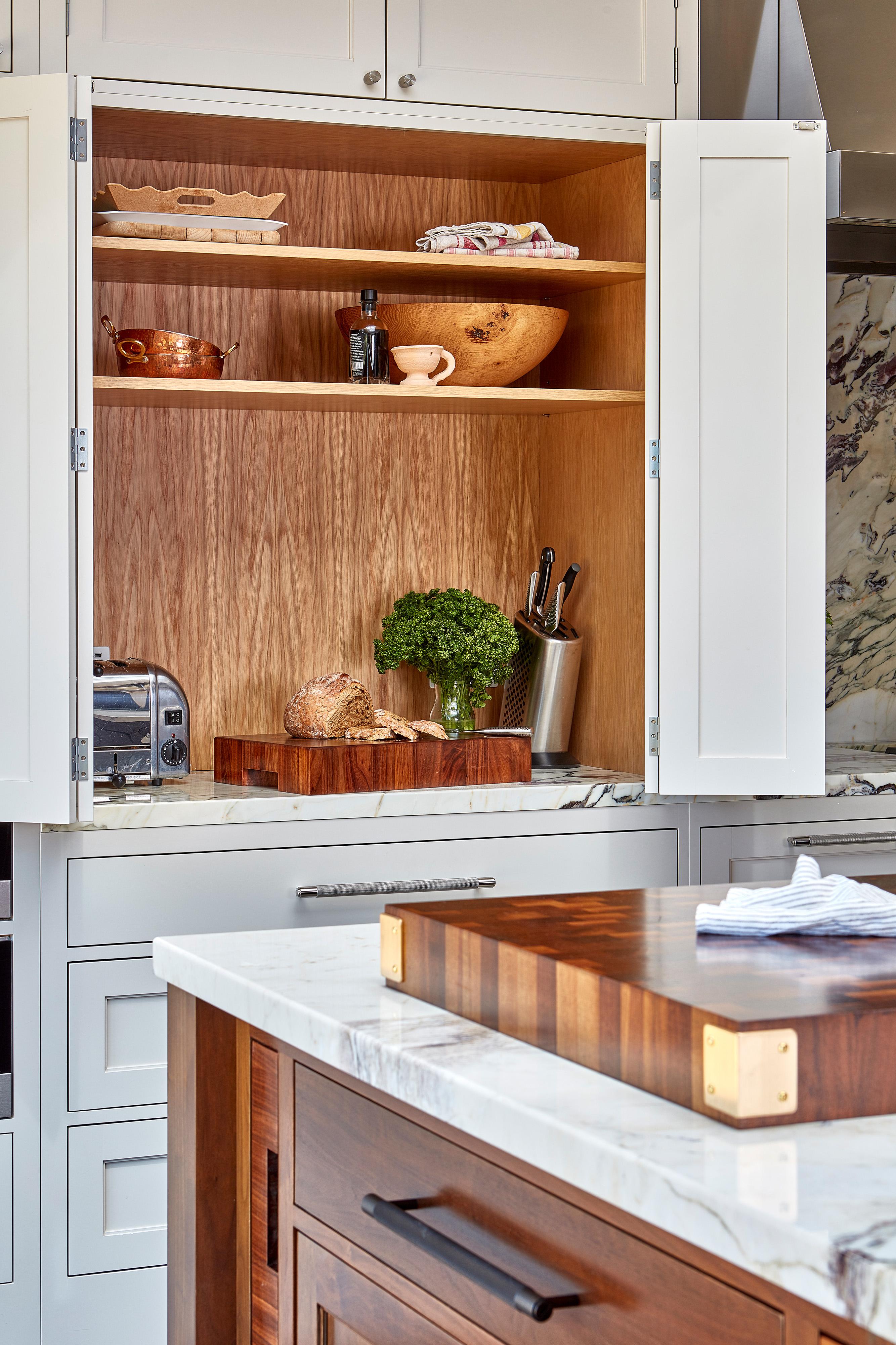 A kitchen with white cabinets and a natural wood interior. Shelves display a copper pot, bowls, a pepper grinder, and a bottle. A chopping block holds parsley and a bread loaf. Nearby are a toaster and knife block. Marble countertops add elegance