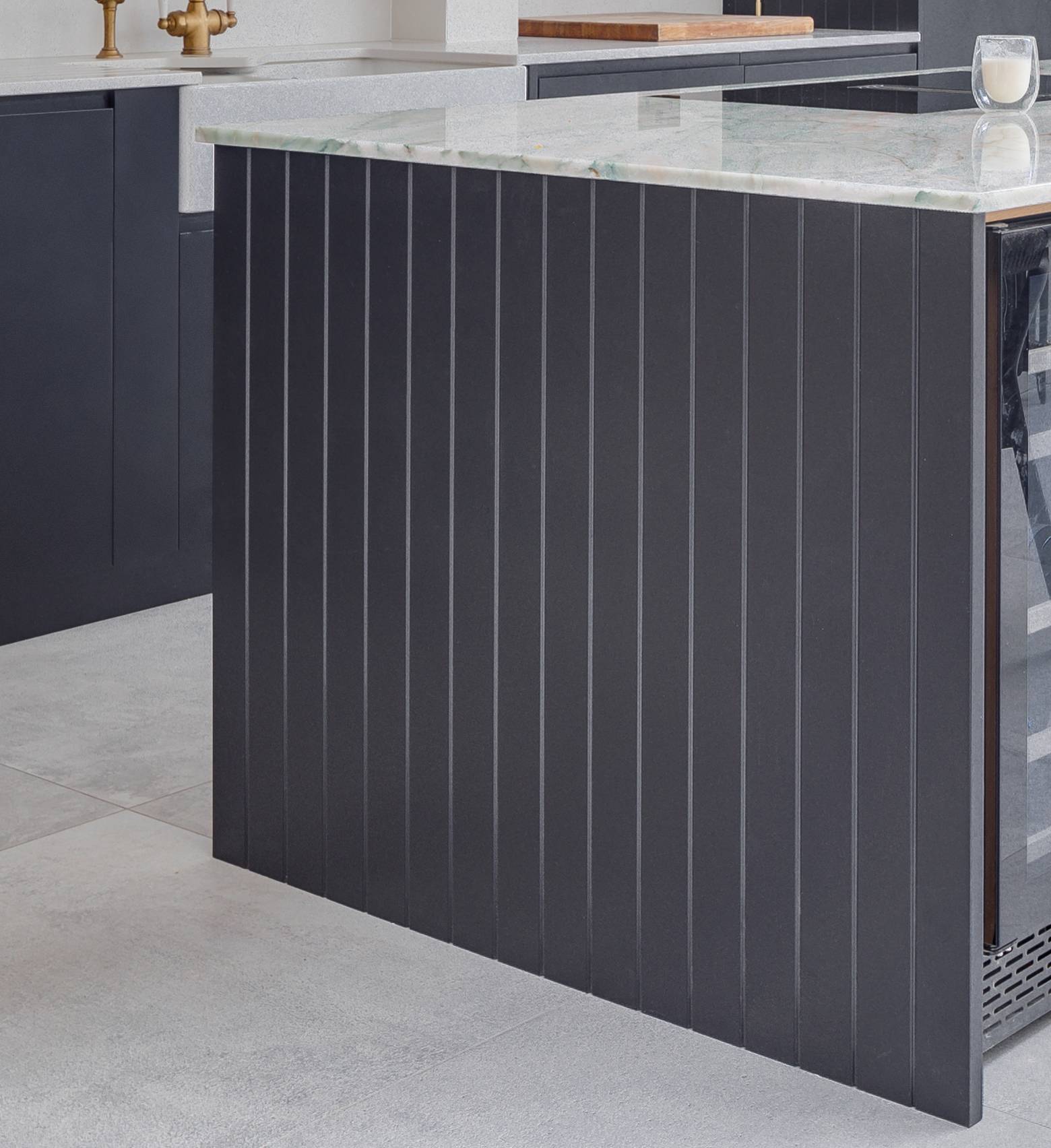 A modern kitchen features a dark vertical panel island with a marble countertop, integrated wine fridge, and light gray floor tiles. A glass of milk sits on the counter.