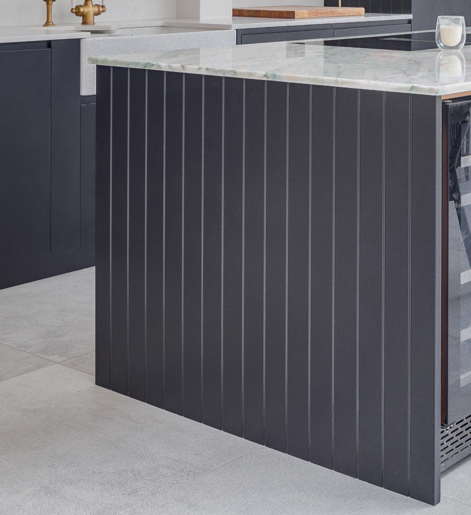 A modern kitchen features a dark vertical panel island with a marble countertop, integrated wine fridge, and light gray floor tiles. A glass of milk sits on the counter.