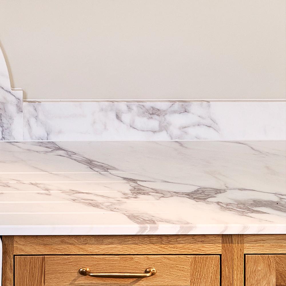 A close-up of a white marble countertop with prominent grey veining, a matching low marble splashback, and light wood kitchen cabinetry with a brass handle below.