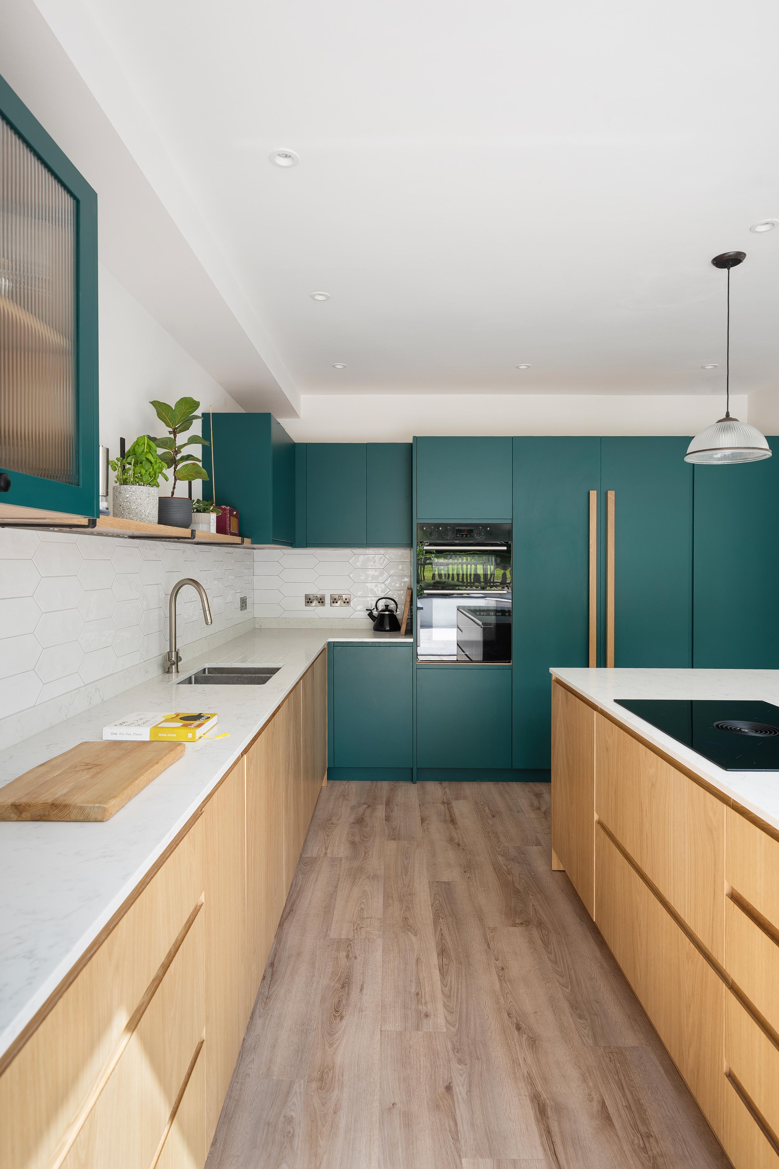 Modern kitchen with teal cabinets and light wood lower cabinets. White countertops and hexagonal tile backsplash. Built-in oven, induction cooktop, and sink. Hanging light fixture and potted plants on shelves. Light wooden floor.