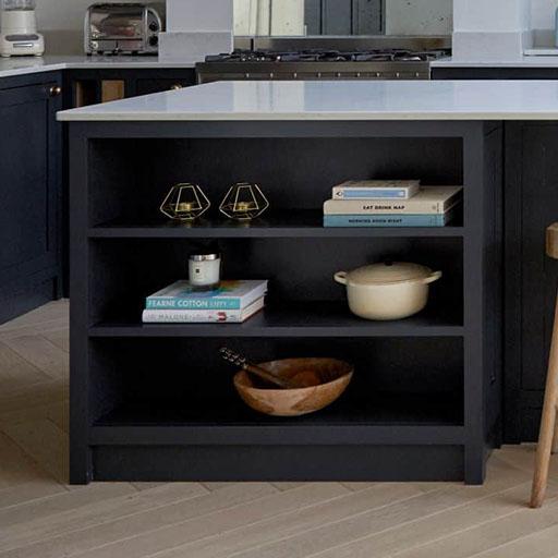 A kitchen island with open shelves displaying books, two glass candle holders, a lidded Dutch oven, a small candle, and a wooden bowl with utensils. The island has a white countertop and dark base.