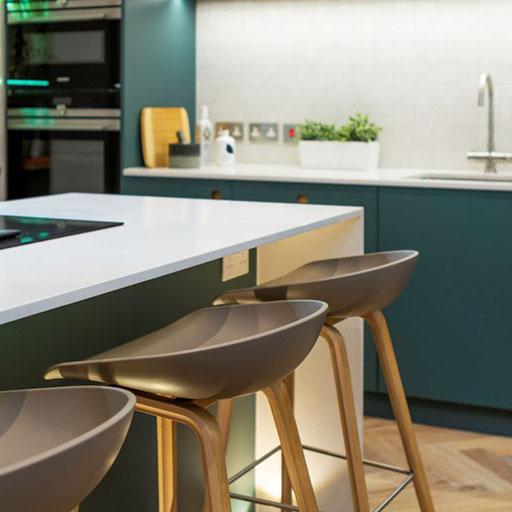 A modern kitchen with teal cabinets, a white island countertop, and three brown barstools with wooden legs. The background shows a sink, potted plant, and built-in appliances.