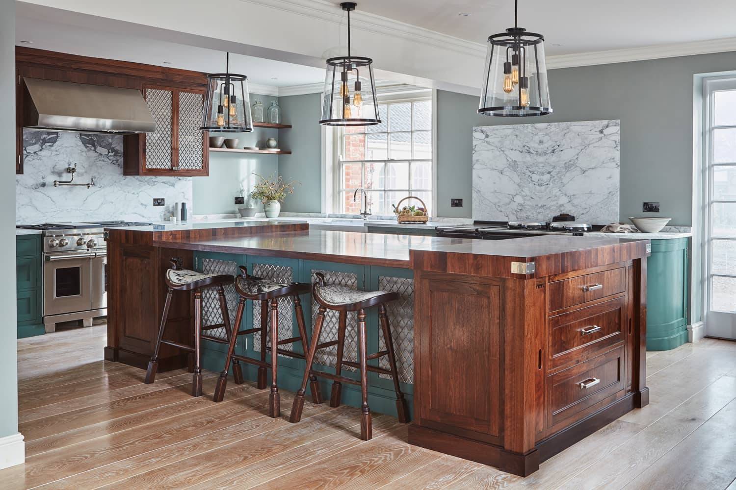 A modern kitchen featuring a large wooden island with four stools. The countertops are marble, and there are two pendant lights above. Appliances are stainless steel, and the cabinets are green with glass fronts. Light wood flooring complements the space.