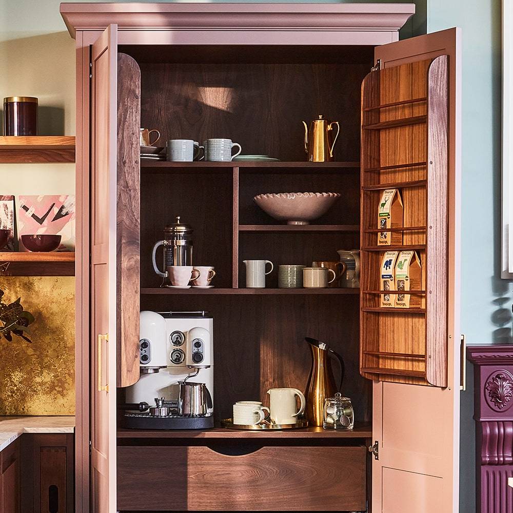 An open wooden cabinet displays a coffee machine, mugs, a French press, teapots, and coffee supplies, neatly arranged on shelves and door racks in a cozy kitchen setting.