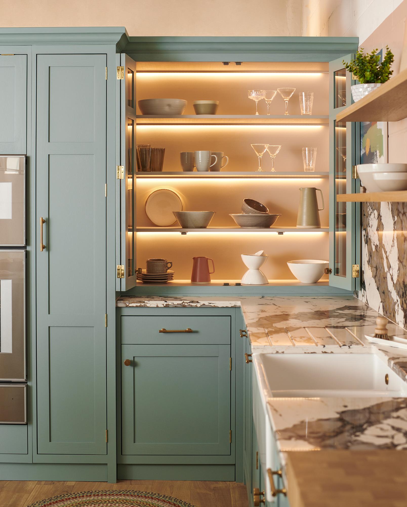 A modern kitchen with sage green cabinets, open shelving displaying neatly arranged dishware and glassware with soft under-cabinet lighting. The countertops are marble, and there's a farmhouse sink with a brass faucet. Shelves hold bowls, mugs, and glasses