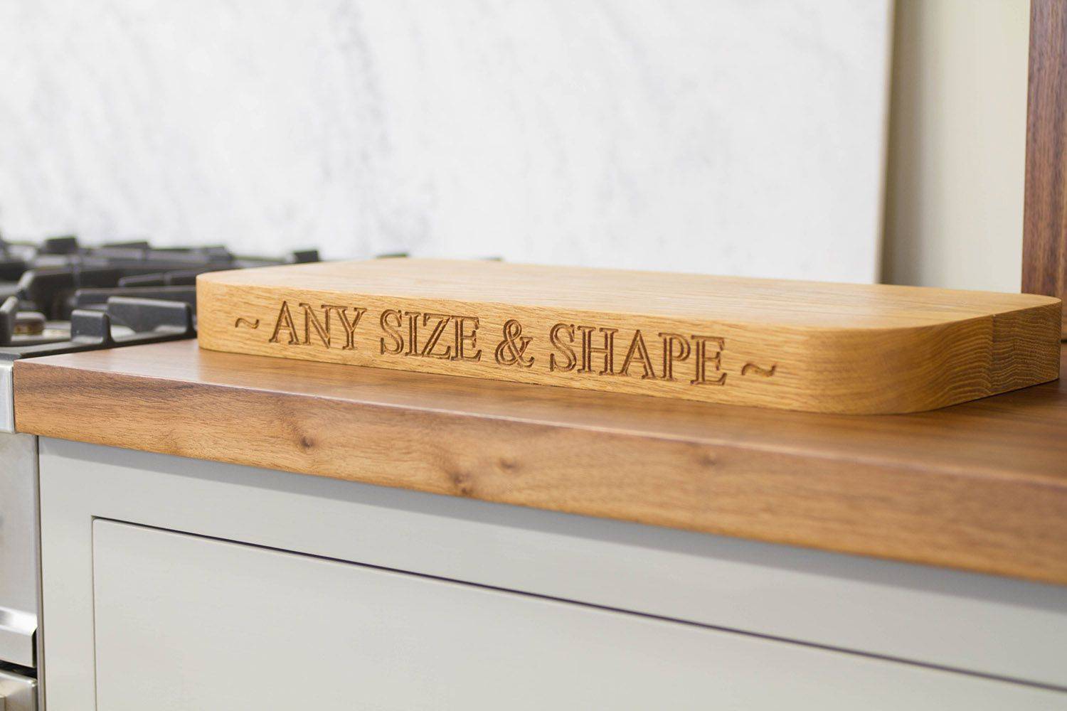 Wooden cutting board on a kitchen counter, engraved with the phrase 'any size & shape'. The counter is next to a stovetop, and marble backsplash partially visible in background.