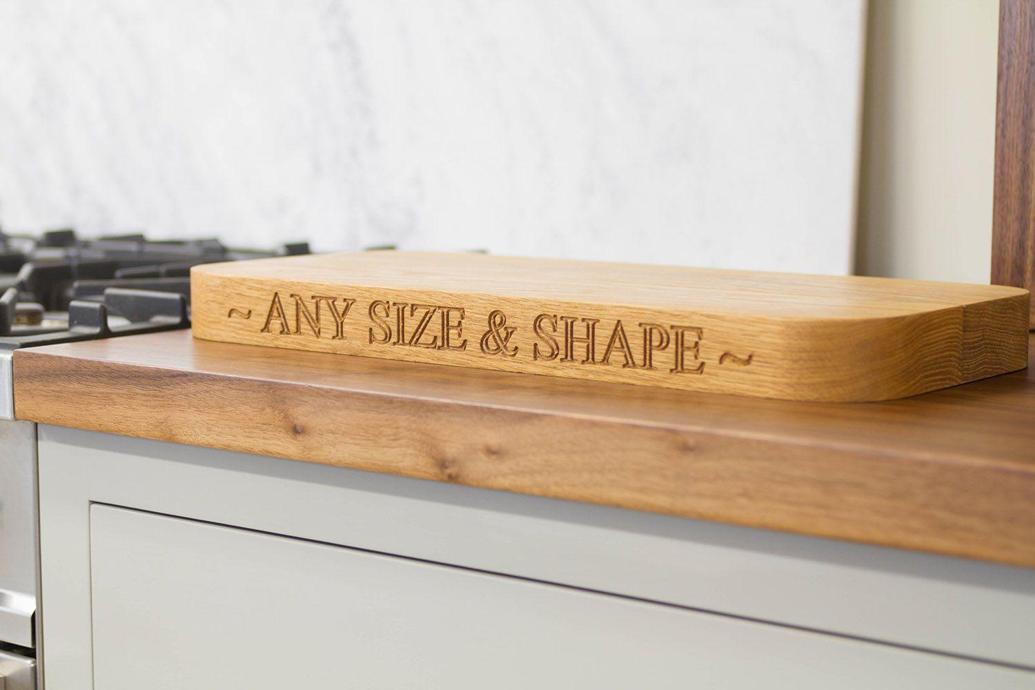 Wooden cutting board on a kitchen counter, engraved with the phrase 'any size & shape'. The counter is next to a stovetop, and marble backsplash partially visible in background.