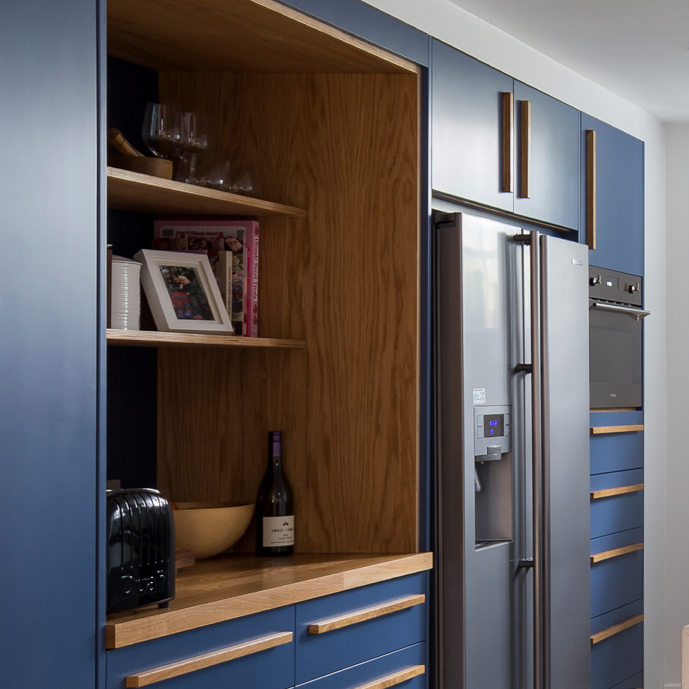 A modern kitchen with blue cabinets, wooden shelves, a stainless steel refrigerator, built-in oven, and a black toaster. The shelves display glasses, books, a framed photo, a bowl, and a wine bottle.
