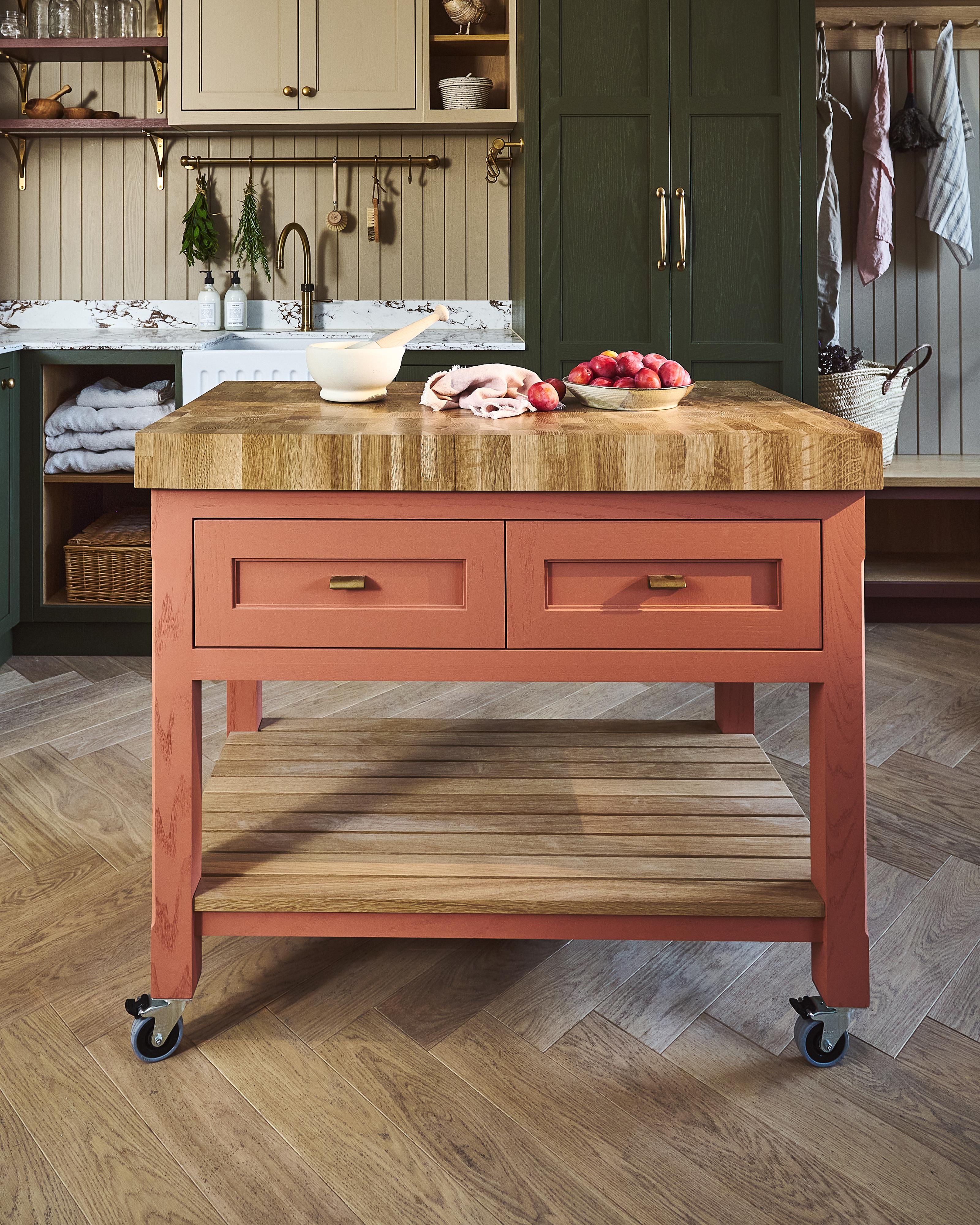 A mobile island with a butcher's block worktop painted in our Pamment colour and stands on wheels. On top are a bowl, a towel, and a pile of radishes. The kitchen has wood floors, green cabinets, and hanging utensils.