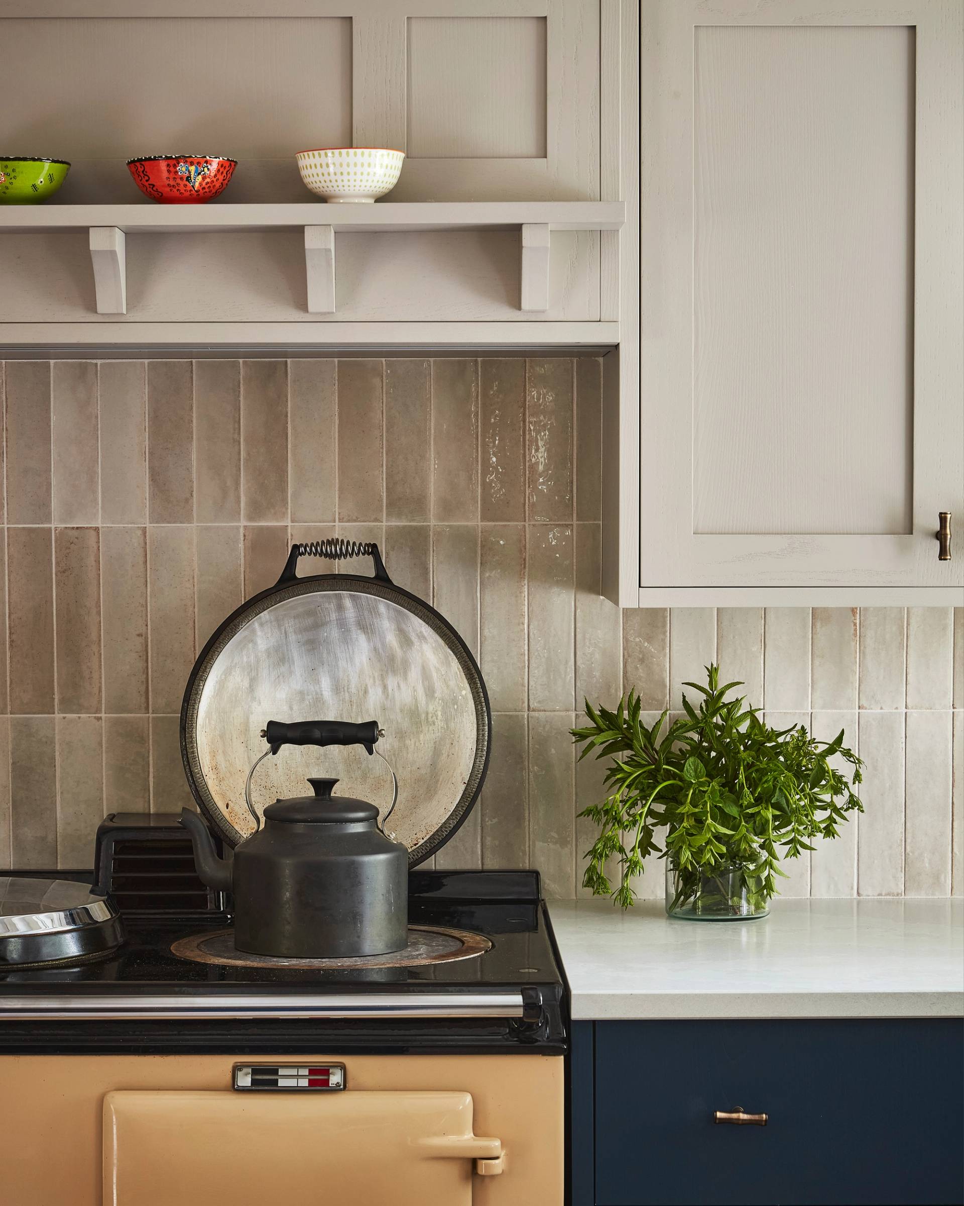 A black kettle sits on a beige stove beneath a lifted metal lid in a cozy kitchen. Above, three patterned bowls rest on a shelf. A glass jar with green herbs decorates the white countertop next to navy cabinets.