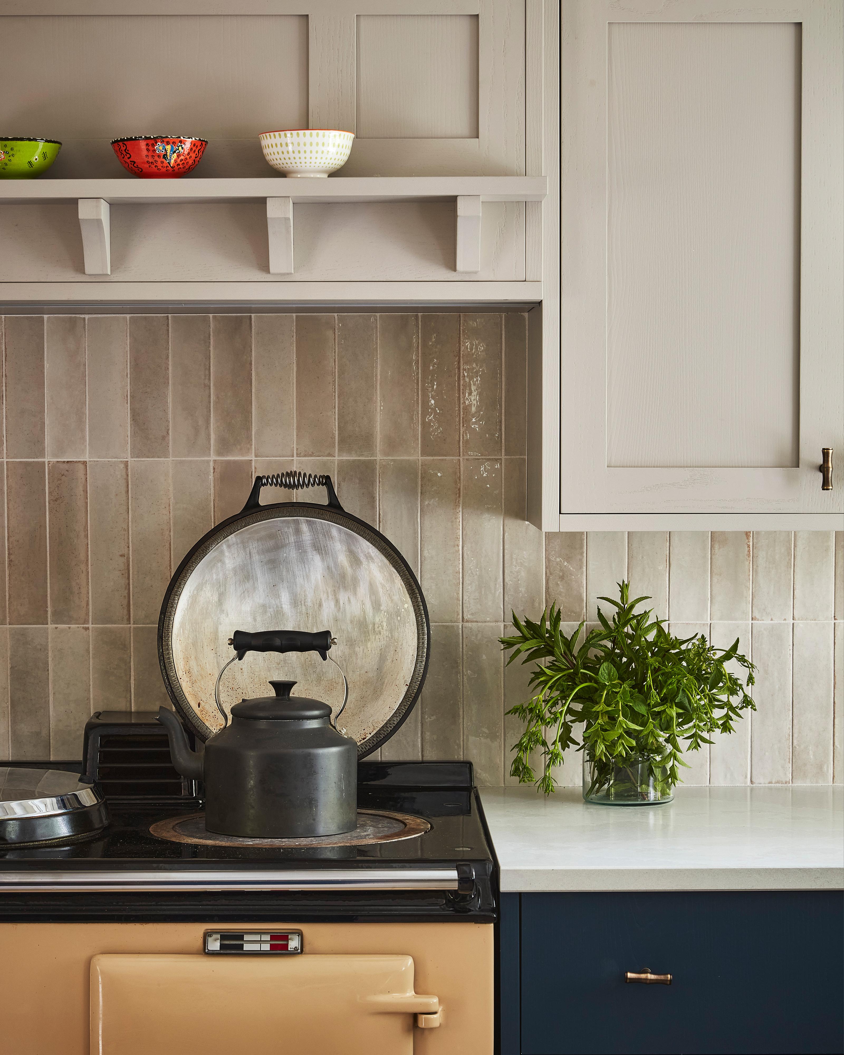 A black kettle sits on a beige stove beneath a lifted metal lid in a cozy kitchen. Above, three patterned bowls rest on a shelf. A glass jar with green herbs decorates the white countertop next to navy cabinets.