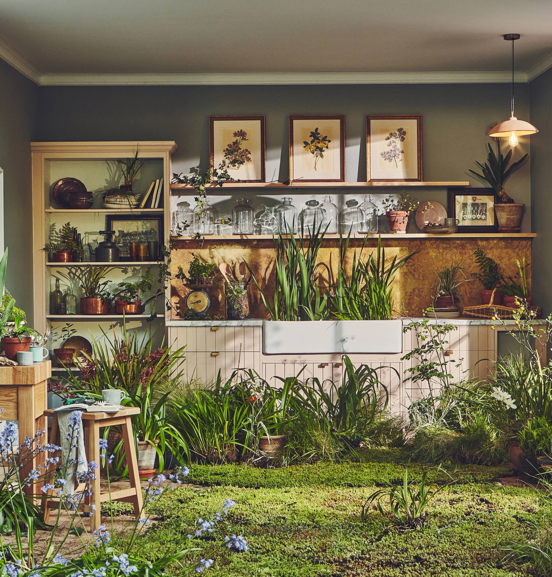 A surreal, indoor garden kitchen where the floor is covered in a lush, green mossy lawn. Tall grasses and potted plants grow directly around a white farmhouse sink and light pink cabinetry. Above the sink, a gold-toned backsplash is topped with wooden shelves holding vintage glass jars and botanical prints. The room is filled with an abundance of greenery, including ferns and flowering plants, blurring the line between a functional kitchen and a greenhouse.