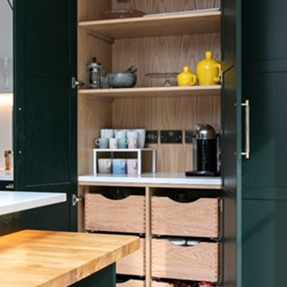 A kitchen cabinet with open dark green doors reveals wooden shelves holding mugs, yellow teapots, a mortar and pestle, a coffee machine, and three drawers with cut-out handles.