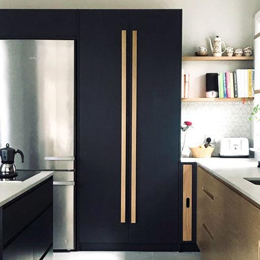 Modern kitchen with black cabinets featuring vertical wood handles, a stainless steel fridge, open shelves with dishes and books, a coffee maker, and light countertops. Natural light brightens the space.