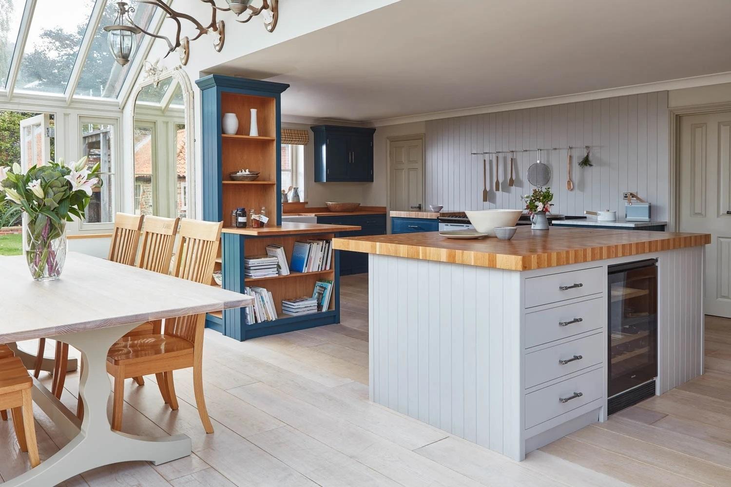 A spacious, sunlit kitchen and dining area featuring a butcher-block island with integrated wine cooler, light oak dining set beneath a glass roof, and navy accent shelving.