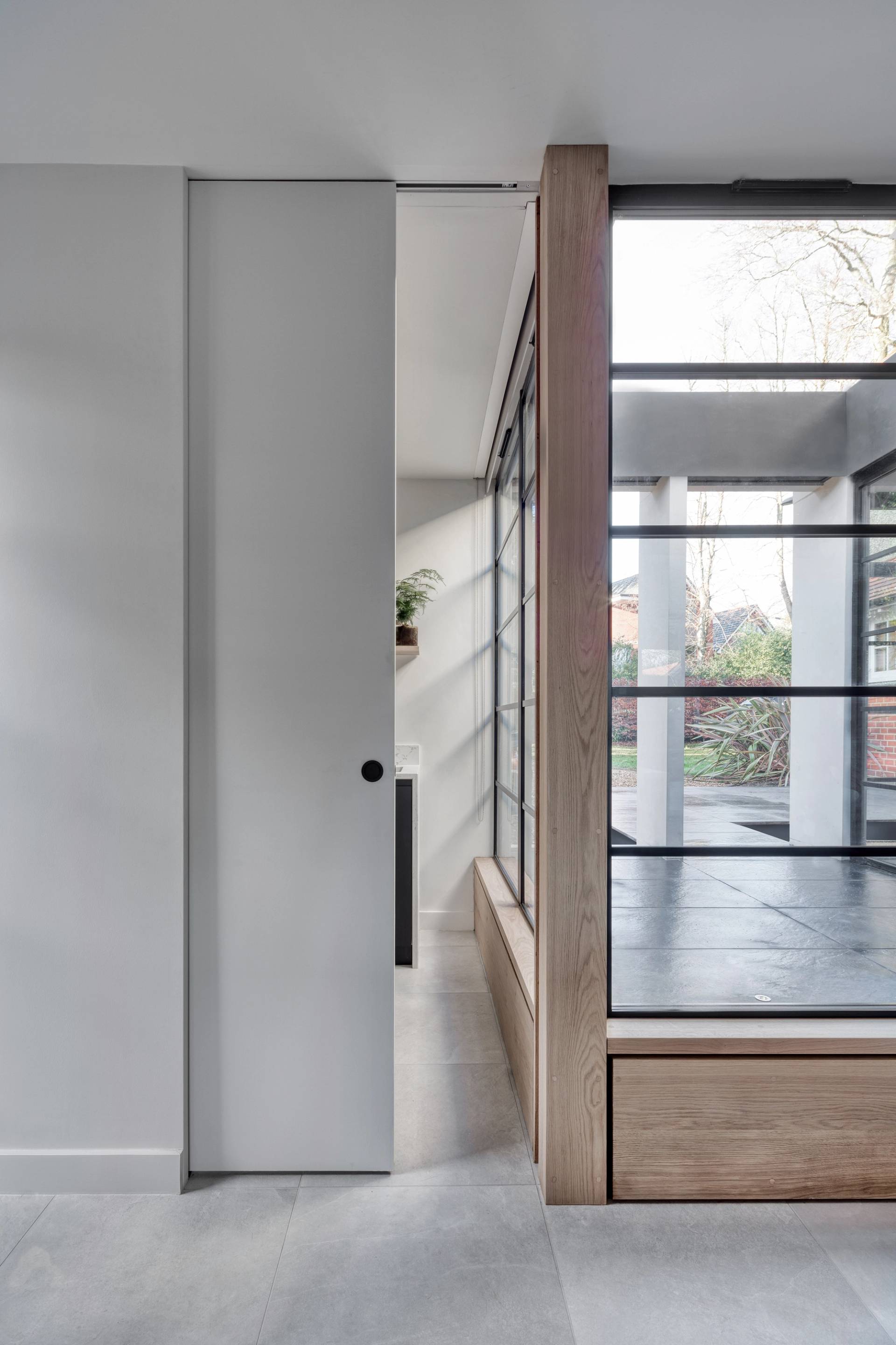 Modern interior showing a sleek kitchen with minimalist black and white design, including a black sink and white countertops. Adjacent to the kitchen is a bright, glass-walled living area with wooden trim, leading out to a garden with green foliage and a brick wall
