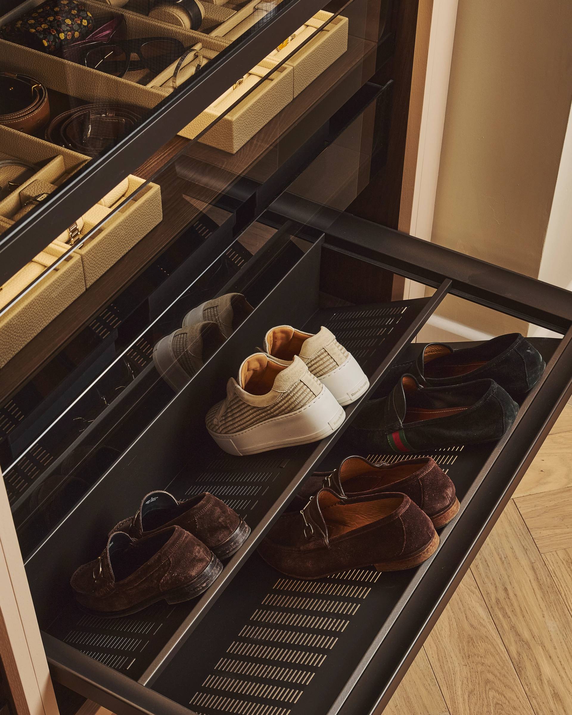 A close-up of a modern closet with open drawers displaying accessories and luxury footwear, including two pairs of sneakers and two pairs of brown loafers neatly arranged on metal shelves.