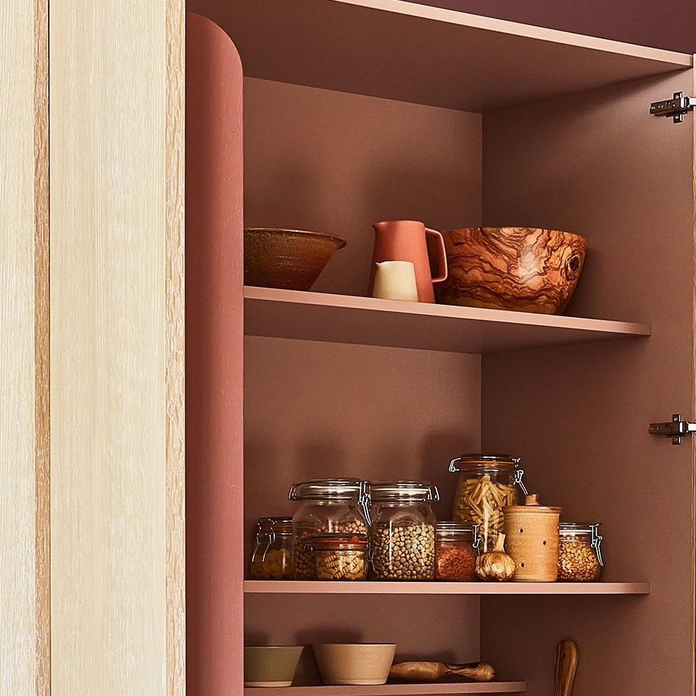 An open kitchen cabinet with brown shelves holds wooden bowls, a ceramic pitcher, glass jars of grains and pasta, and other small kitchen containers against a muted terracotta background.