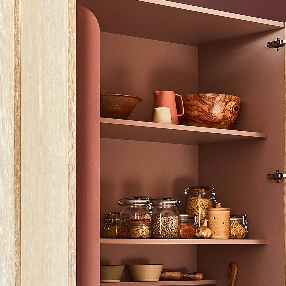 An open kitchen cabinet with brown shelves holds wooden bowls, a ceramic pitcher, glass jars of grains and pasta, and other small kitchen containers against a muted terracotta background.