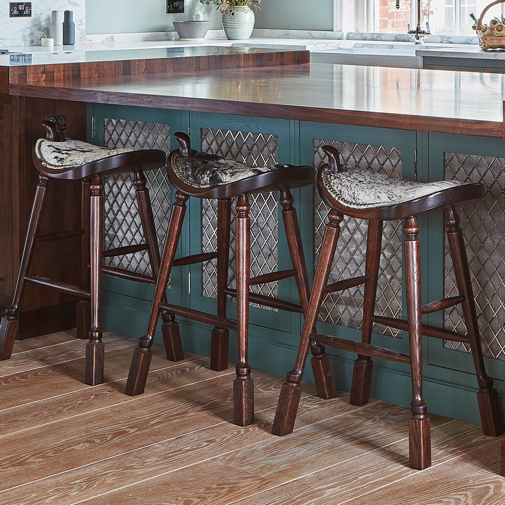 Three wooden barstools with upholstered seats are lined up at a kitchen island with a dark wood countertop and patterned cabinet panels. The floor is wooden, and the background includes a vase with flowers.
